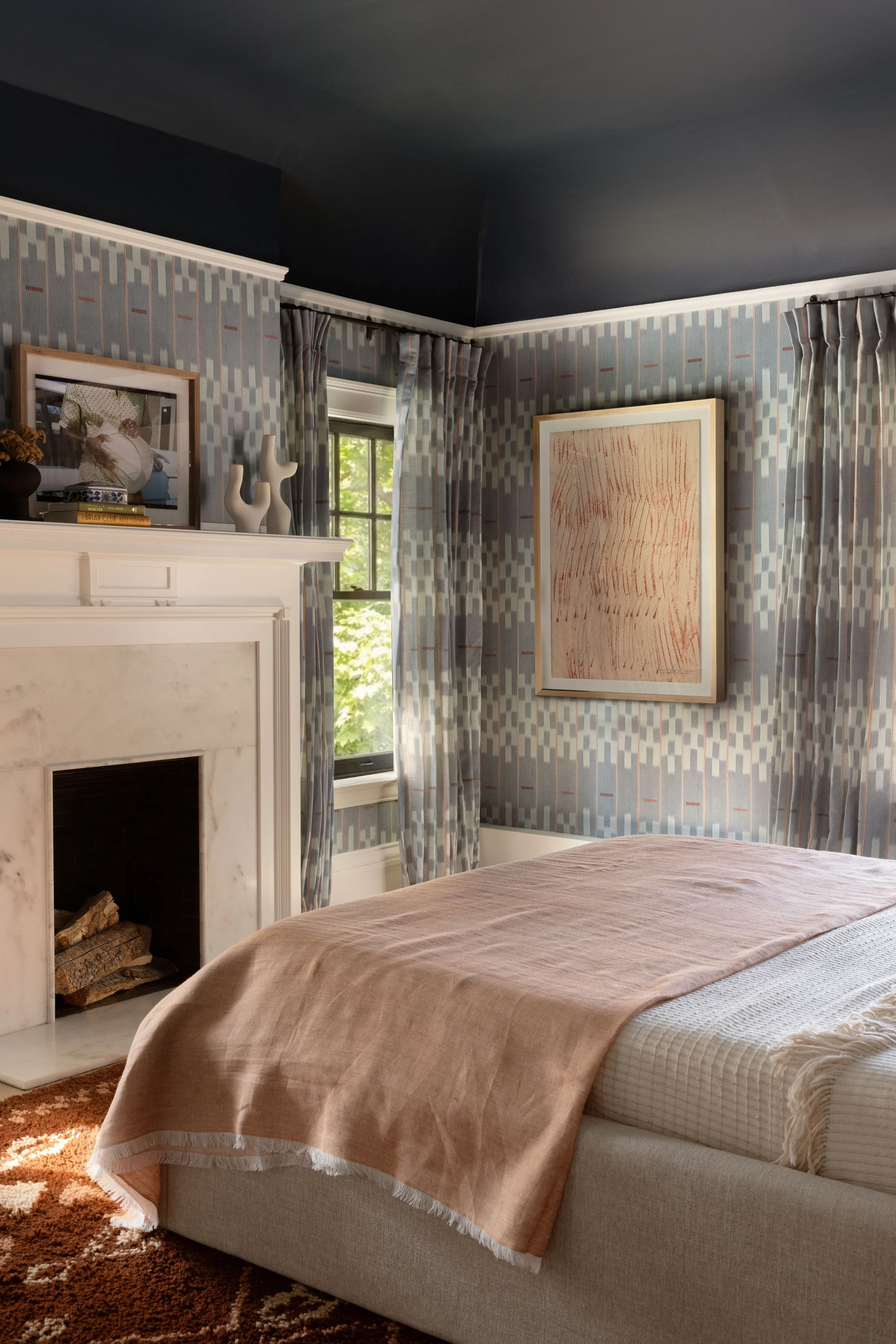 A bedroom with a white fireplace, patterned wallpaper, framed artwork, curtains, and a bed covered with a beige blanket.