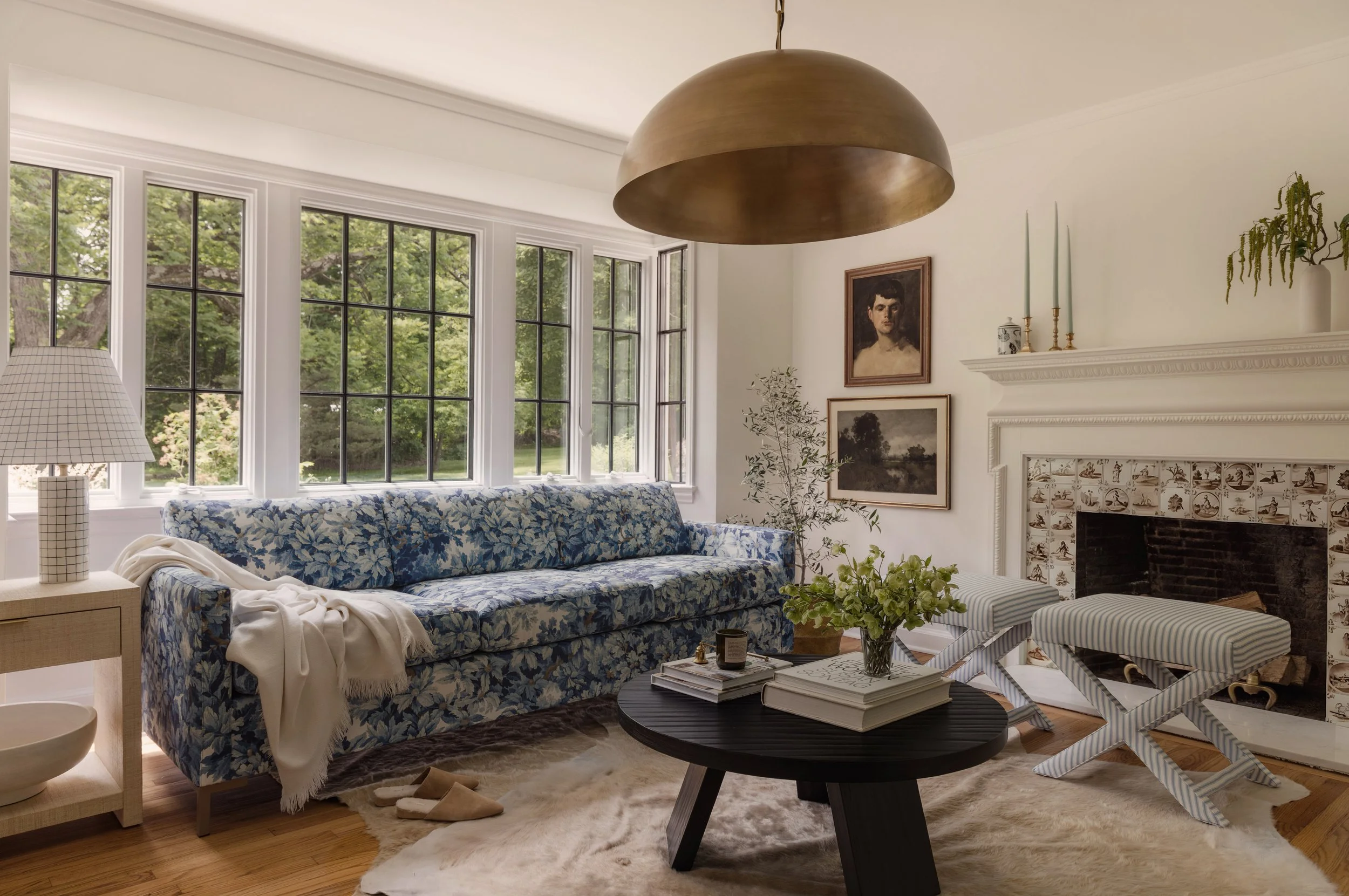 Living room with a large window, blue floral sofa, black coffee table with books and plants, striped ottomans near a white fireplace, and a gold dome pendant light hanging from the ceiling.