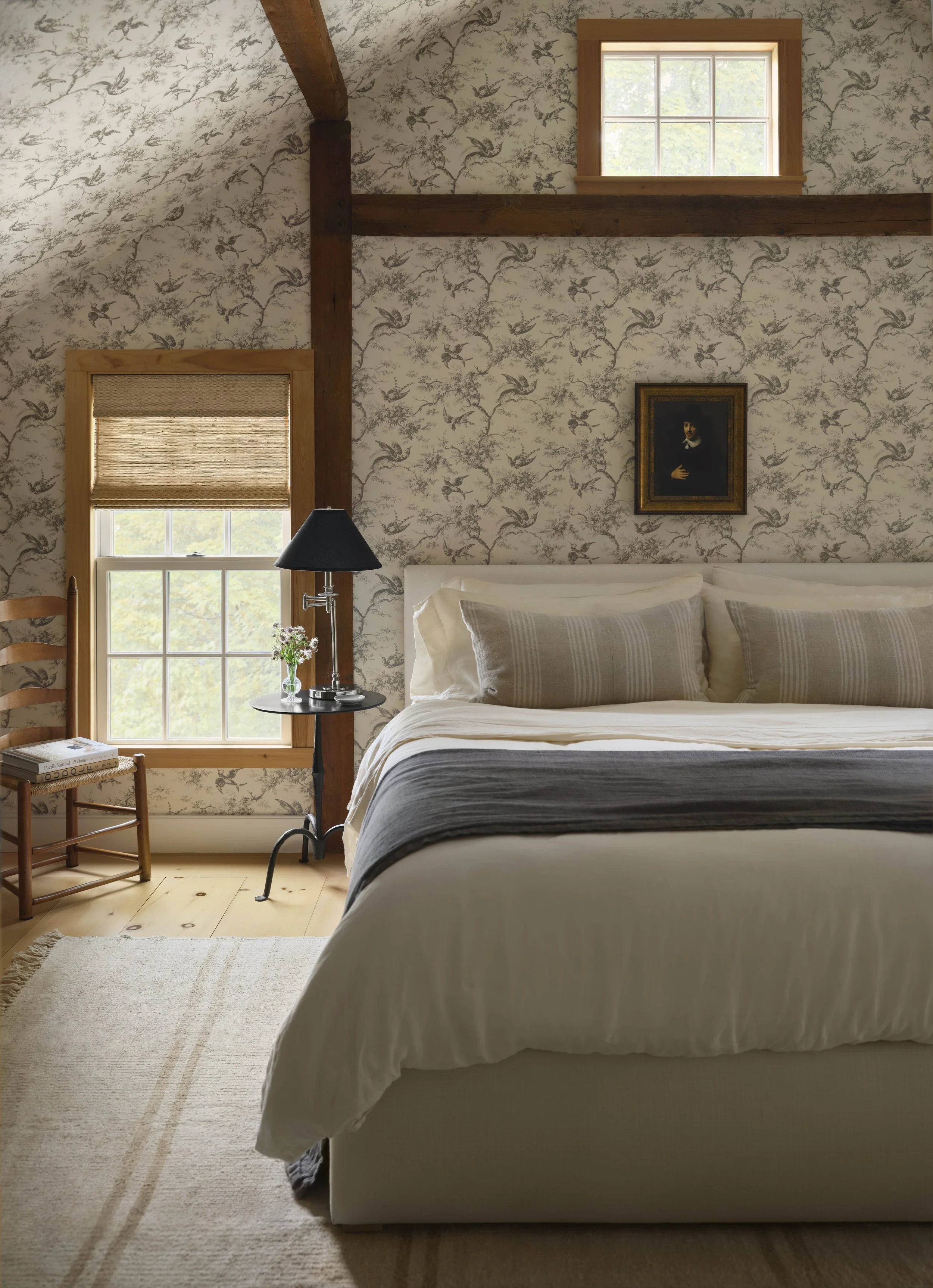 Westchester and Fairfield County interior designer creates a bedroom with floral wallpaper, a bed with white bedding and pillows, a small round black side table with a lamp and flowers, a wooden chair with a book, and a framed portrait on the wall.