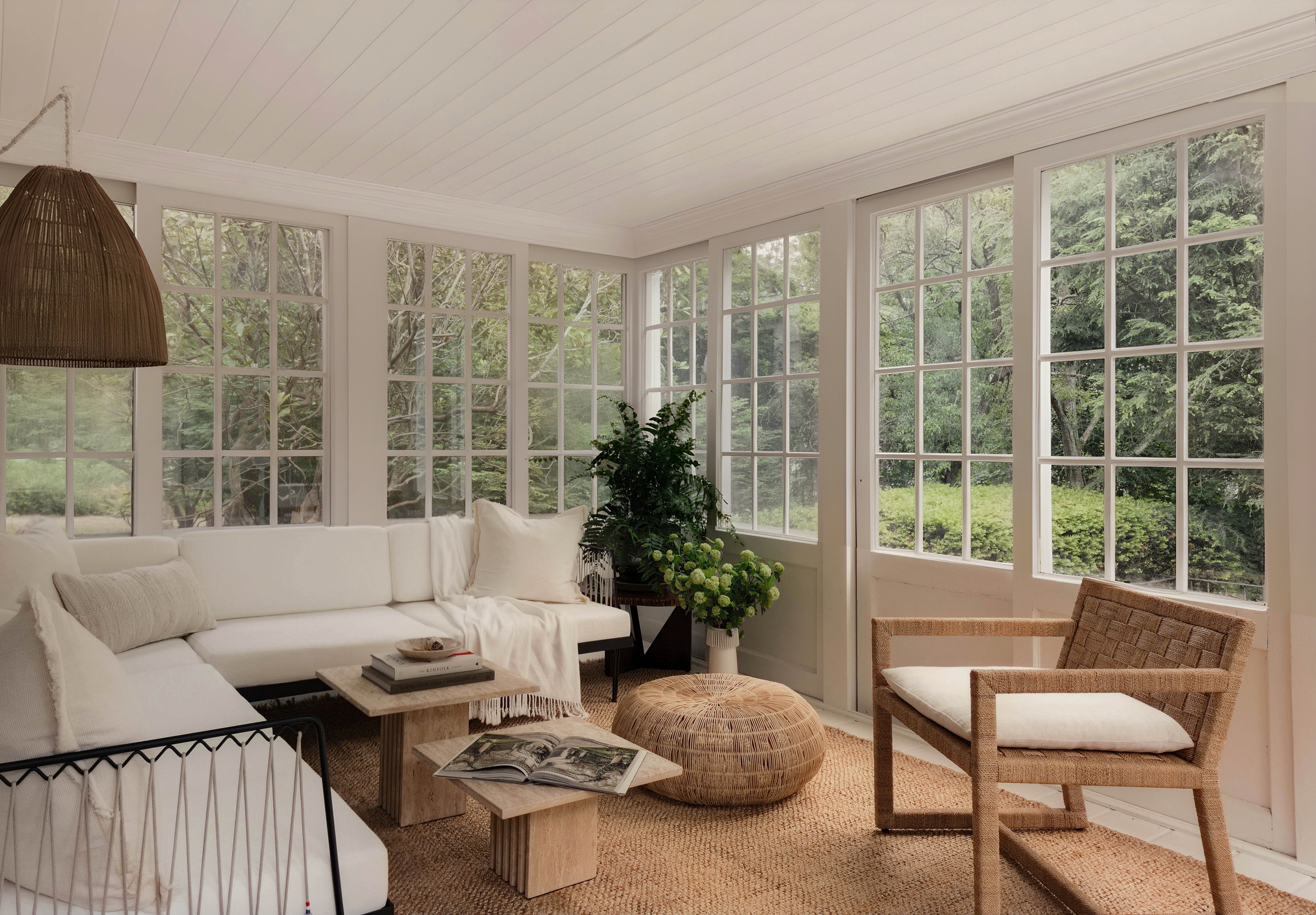 Bright sunroom with white sofa, wicker pouf, armchair, potted plants, surrounded by large windows showing greenery outside, decorated in a natural, minimalist style.
