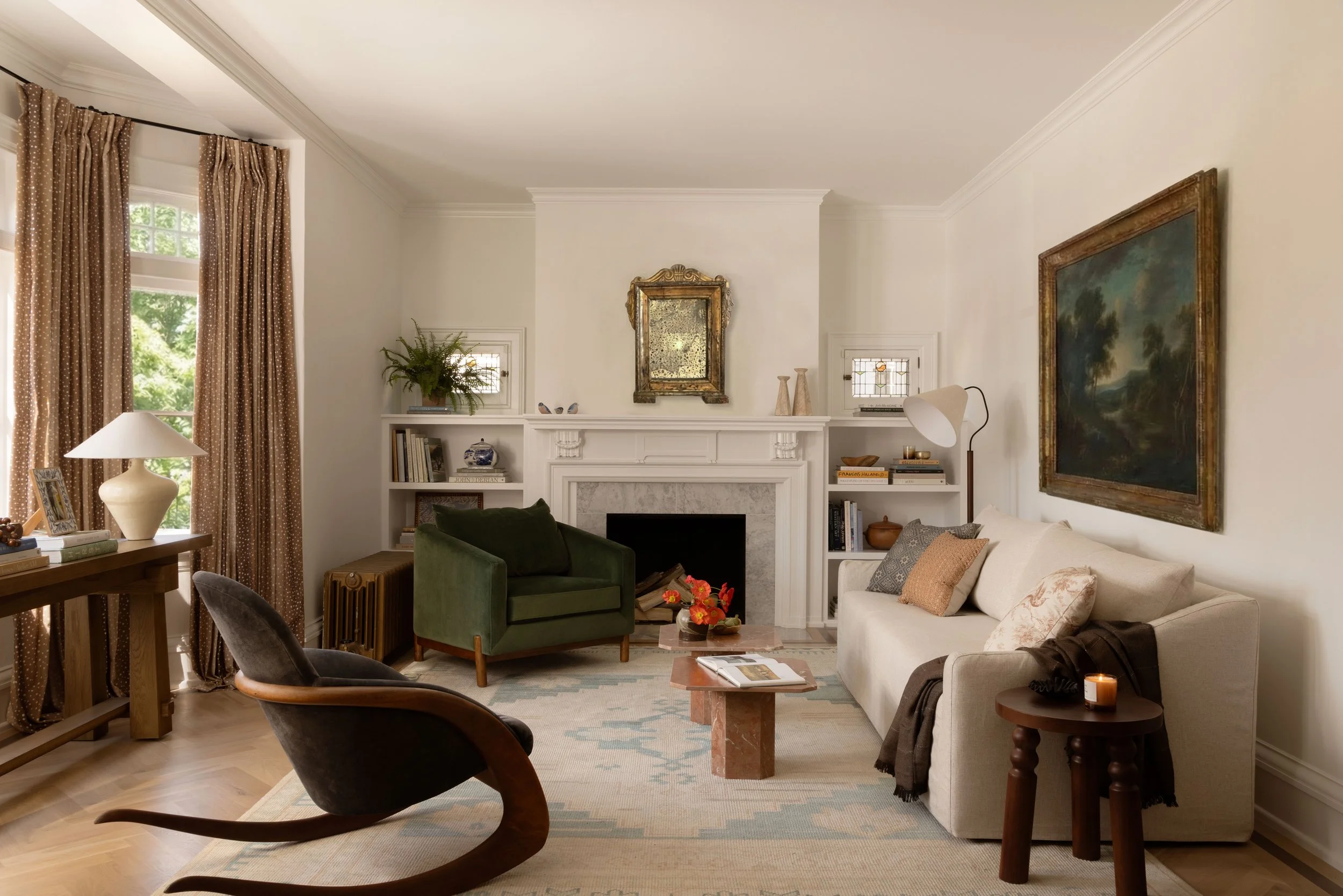 Living room with white walls, a fireplace, beige sofa with pillows, a green armchair, a black rocking chair, a wooden side table with a candle, bookshelves, a large flat-screen TV, and patterned curtains.