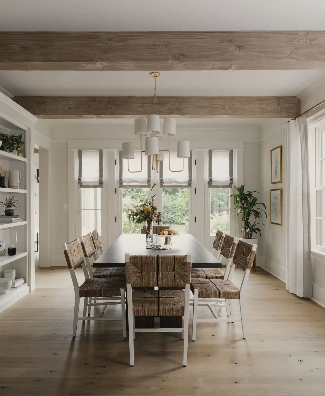 Bright dining room with a large black table, surrounded by chairs with striped brown cushions, white frames, and a white chair at each end. A bouquet of flowers and a tray of food are on the table. White shelves with potted plants and books are on th