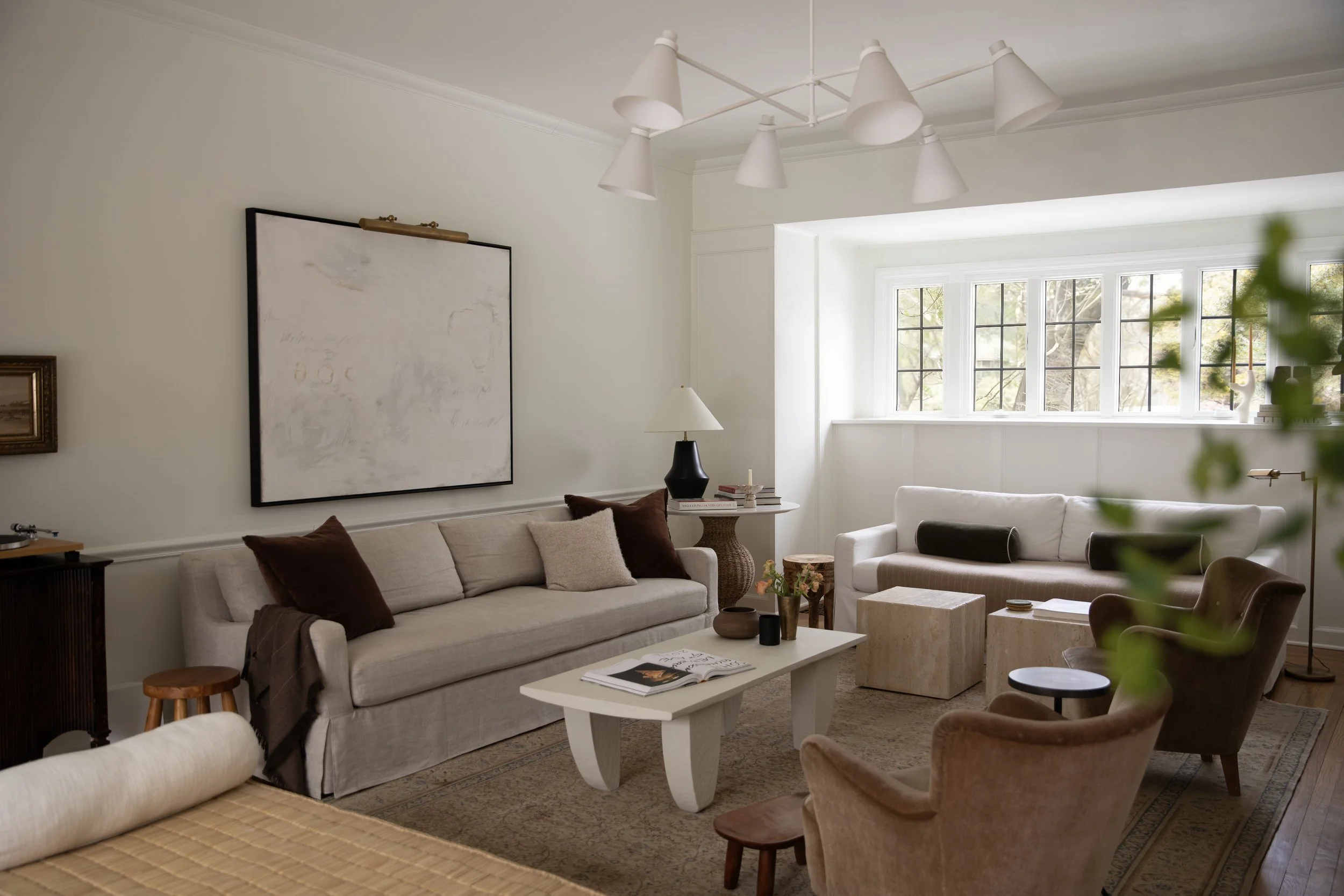 Living room with white sofas, brown and beige armchairs, white coffee table with a vase of red tulips, large window with multiple panes, and various decorative items including lamps and artwork.