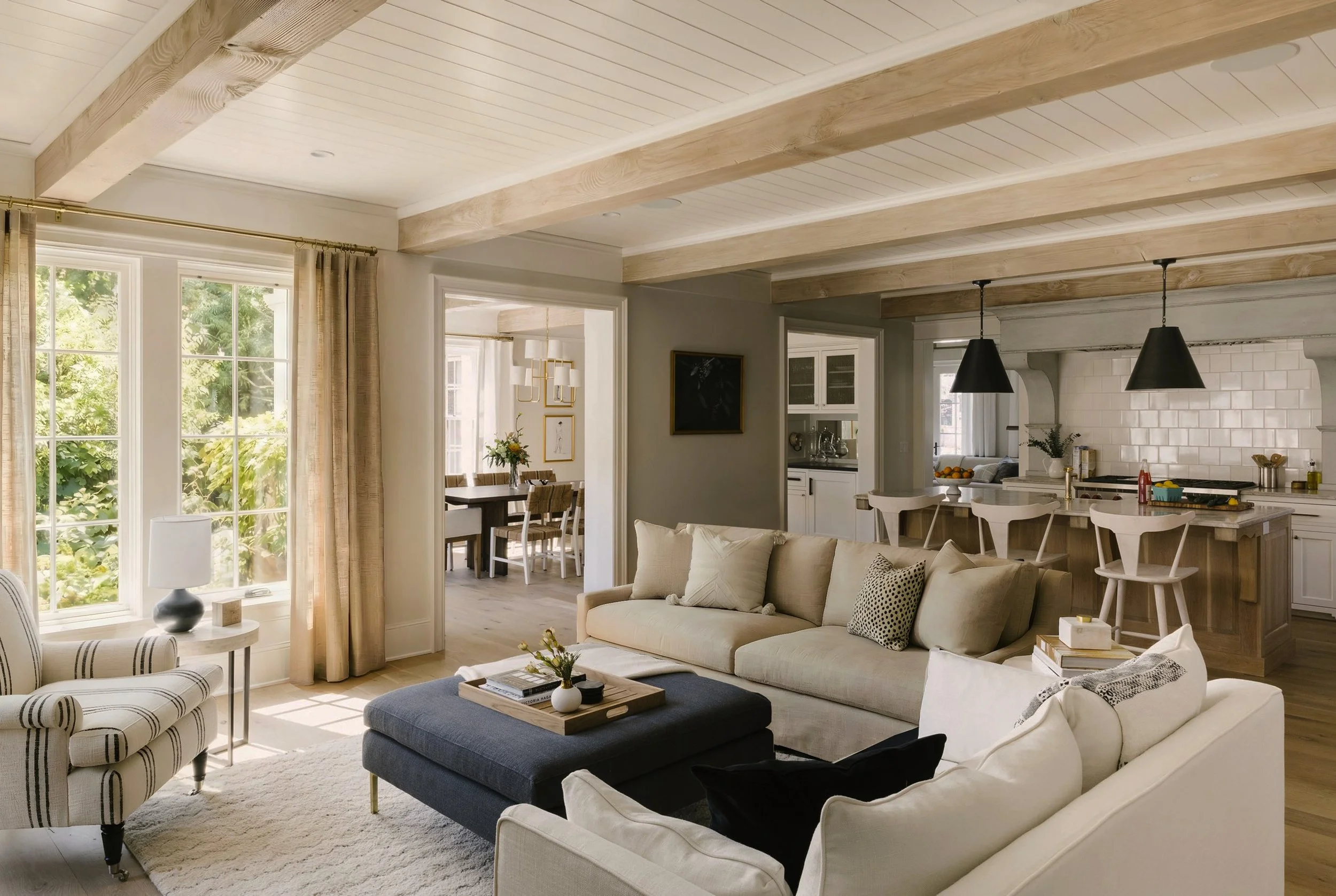 A cozy living room with a black sofa and white armchair, a plaid armchair, a beige rug, and a coffee table with decorative items. The open space includes a view into a dining area and a kitchen with pendant lights, white cabinets, and a tiled backspl