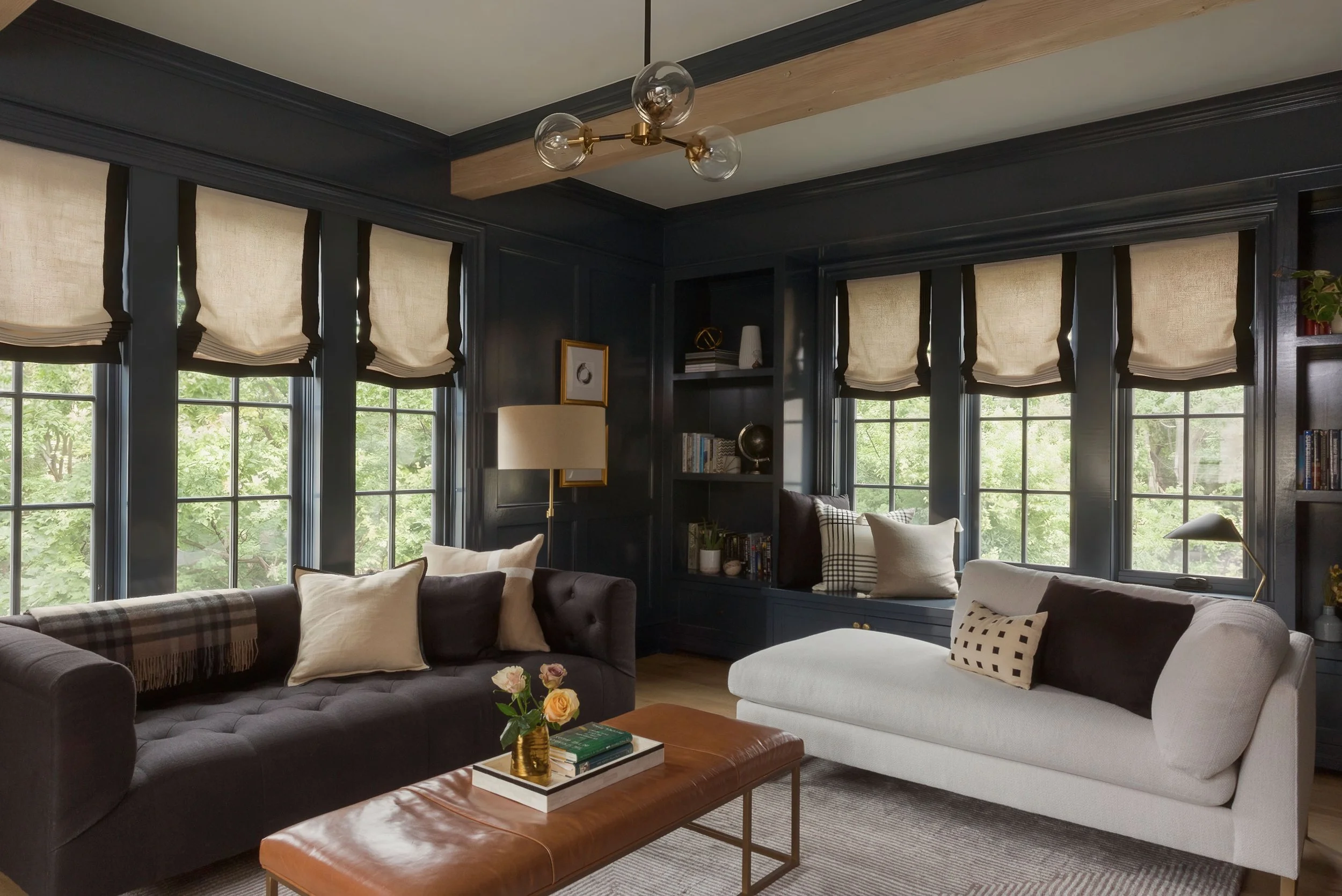 Living room with dark navy walls, multiple windows with beige Roman shades, a black tufted sofa with cream pillows, a white sofa with patterned cushions, a brown leather ottoman with a vase of flowers and books, a floor lamp, built-in shelving with b