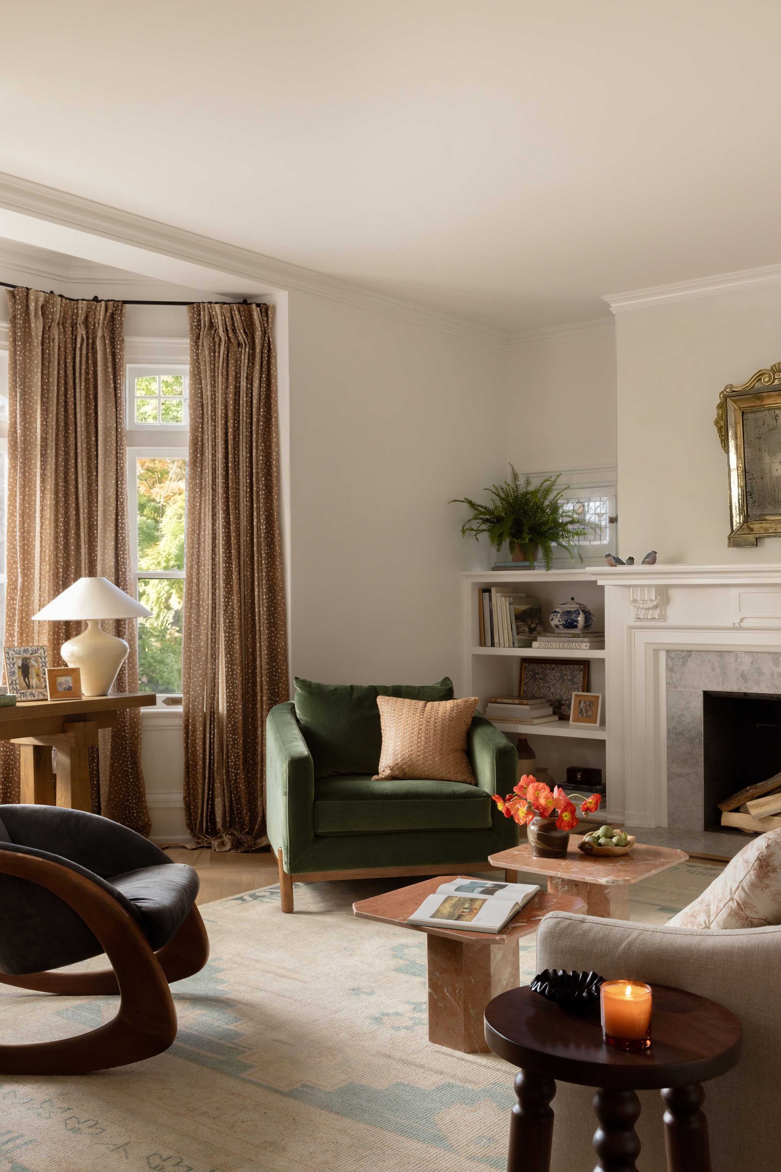 A cozy living room with a green armchair, a fireplace, a window with brown patterned curtains, a side table with a white lamp, a coffee table with a flower vase, and bookshelves with decorative items.