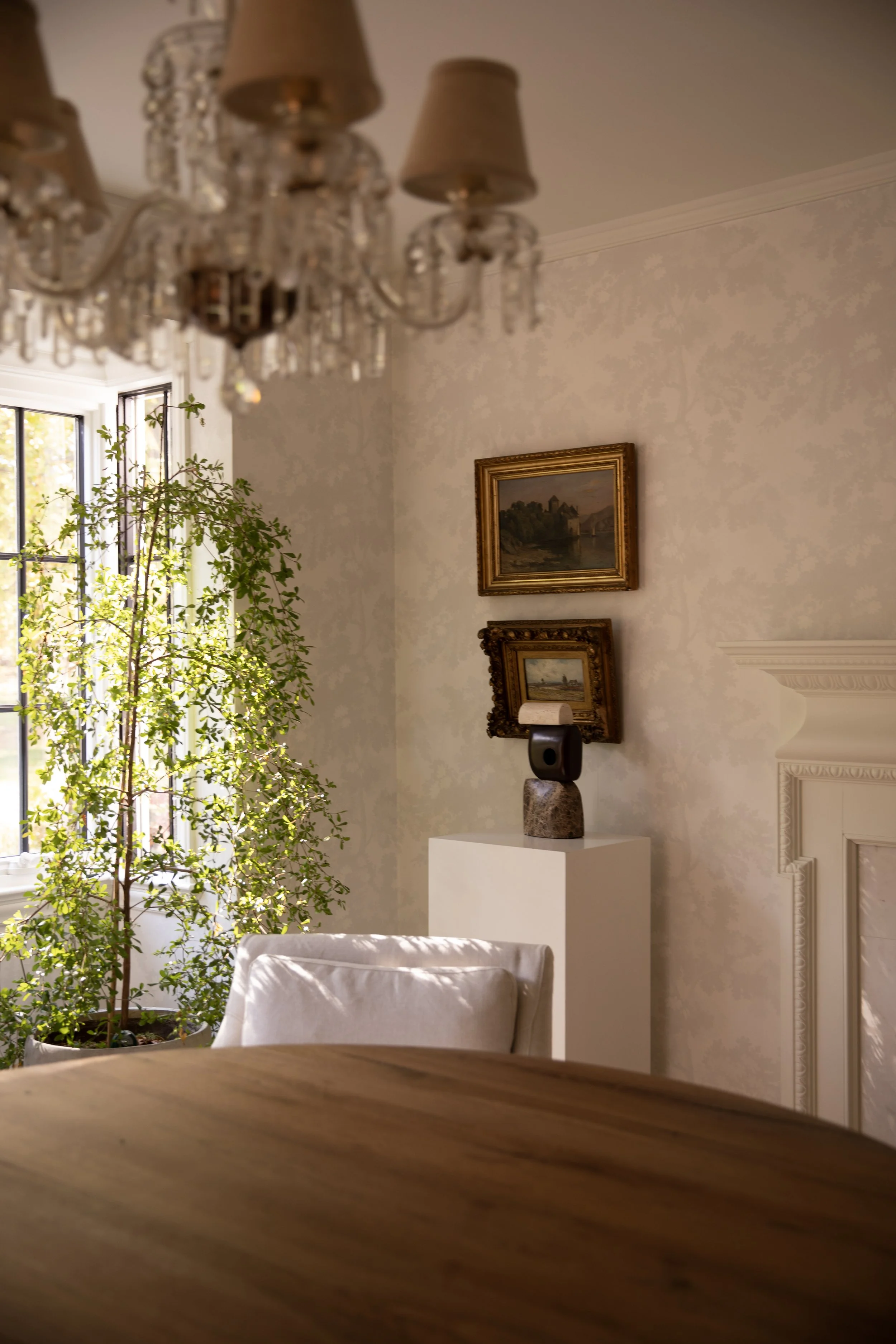 Dining room vignette with vintage crystal chandelier, antique oil painting artwork, a sculptural object on a pedestal and a Shady Lady black olive tree. Subtle wallpaper and a fireplace