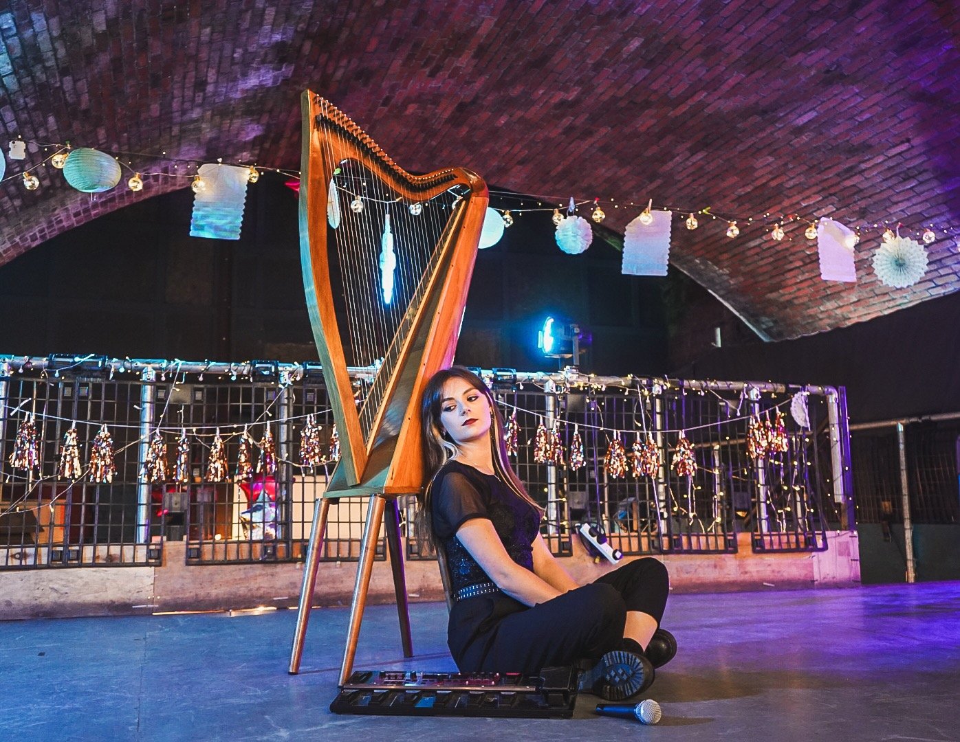Dara sitting on a stage floor, leaning against her harp, with her loop pedal and microphone beside her