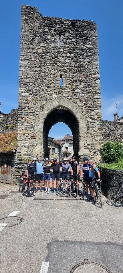 Een groep mensen met fietsen poseert voor een oude stenen stadsmuur met een grote poort, onder een heldere blauwe hemel.