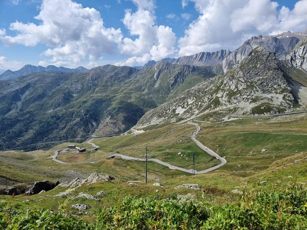 Sierra mamora met groene grasvlaktes, een kronkelende bergweg, enkele kleine gebouwen en bergen op de achtergrond onder een bewolkte hemel.