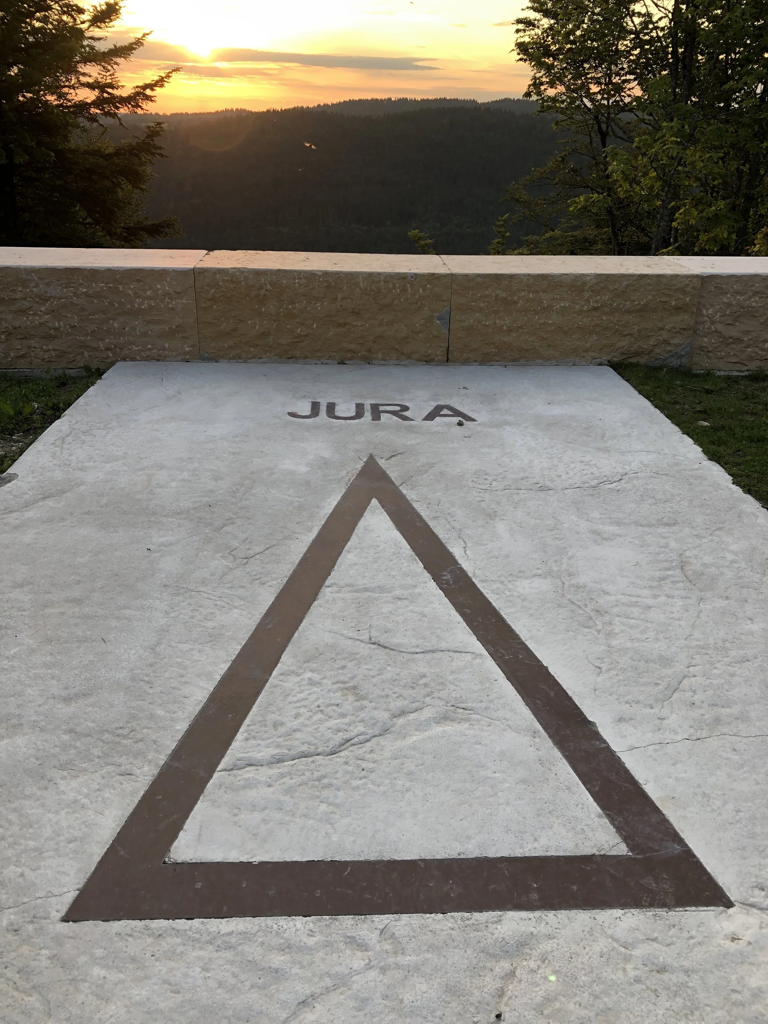 Een indrukwekkend uitzicht op een zonsondergang over een bergketen, met een hellende witte steen met een grote formule. Op de steen staat de tekst 'JURA' en een grote driehoek met een omtrek in bruin.