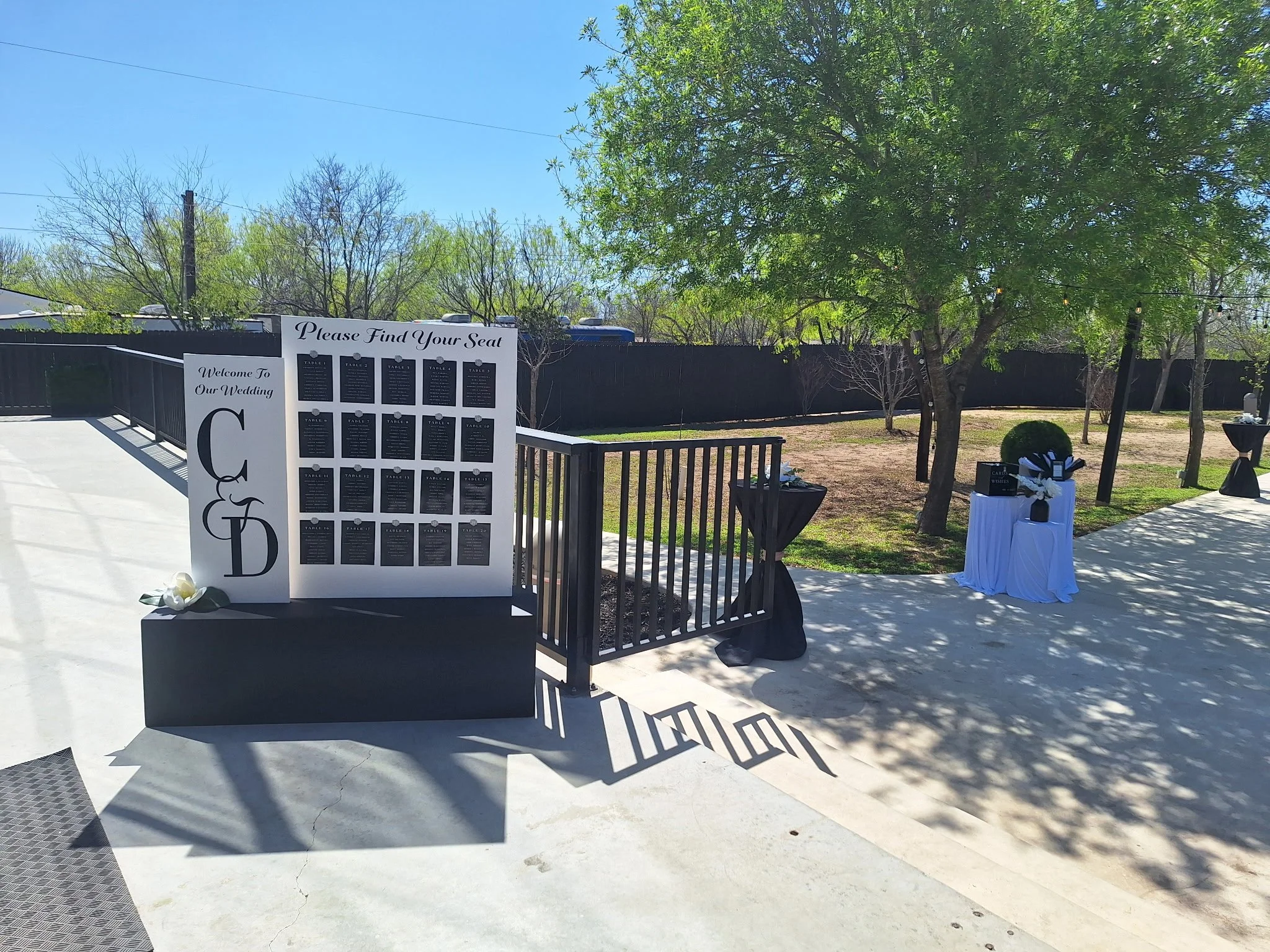 Wedding seating chart display outdoors with black and white decorations, a sign reading 'Please Find Your Seat', and trees casting shadows on the ground.