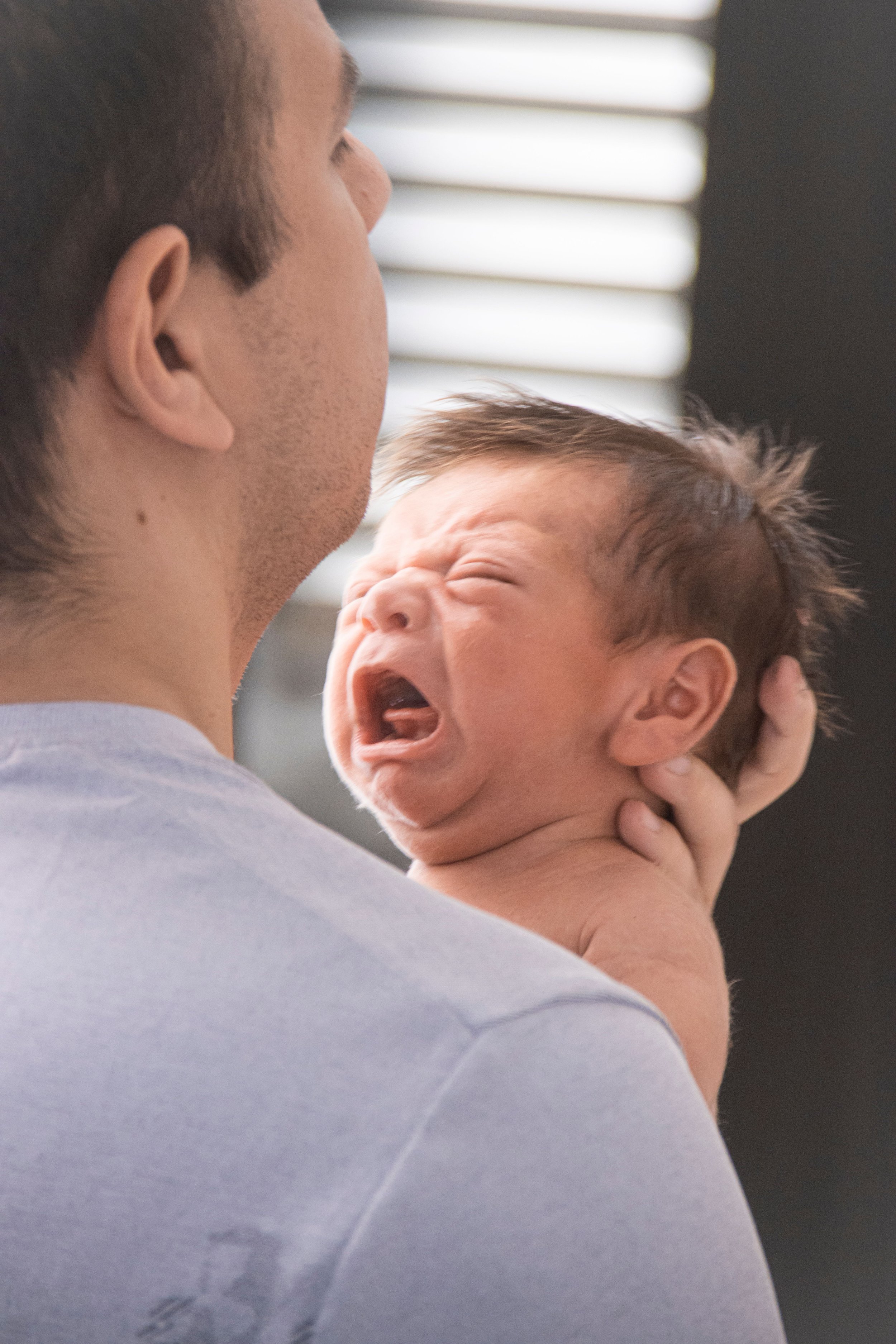 An exhausted parent holding his colicky newborn baby.  A postpartum doula is nearby helping him learn soothing techniques for crying babies and giving him newborn colic relief.
