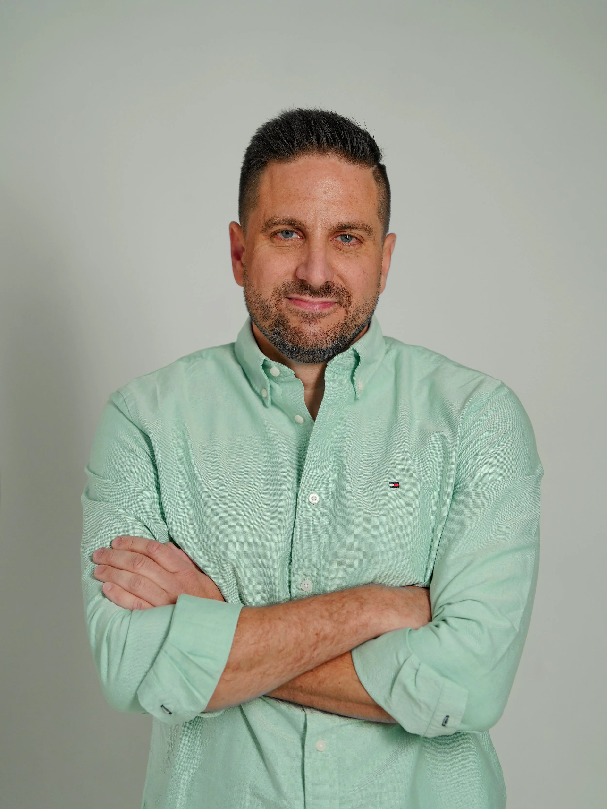 A man with short dark hair, a beard, and blue eyes, wearing a light green button-up shirt with the sleeves rolled up, standing with arms crossed against a plain light background.