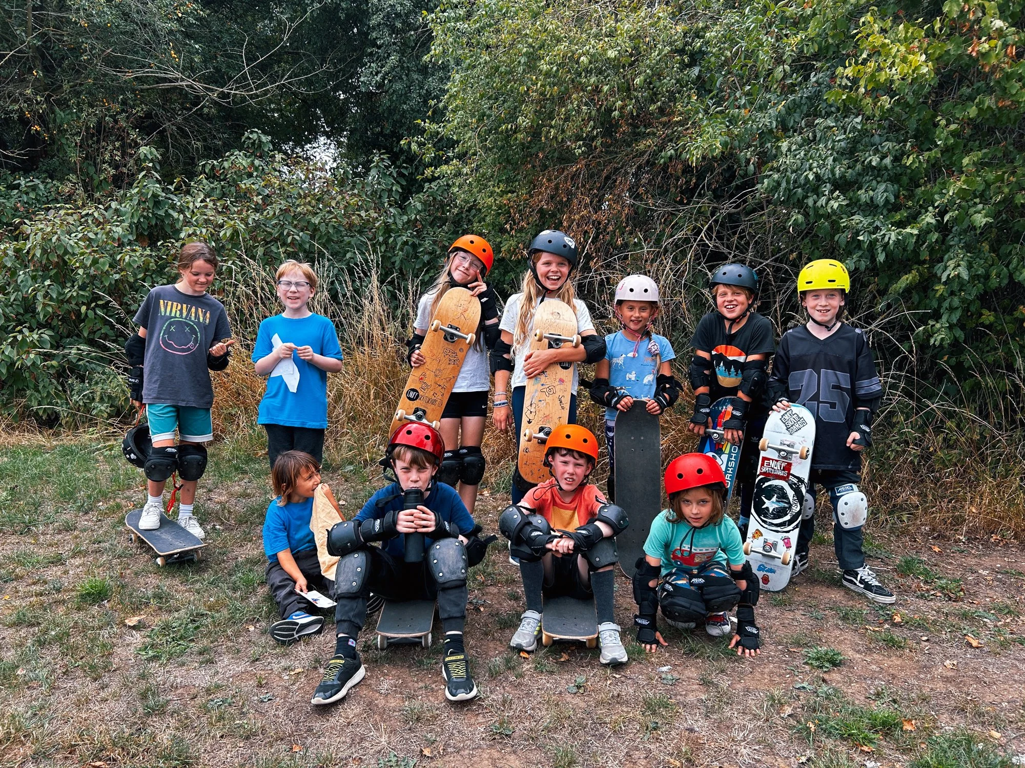 Group of children with skateboards and helmets outdoors, posing for a photo with some standing and others sitting on skateboards, in front of greenery.