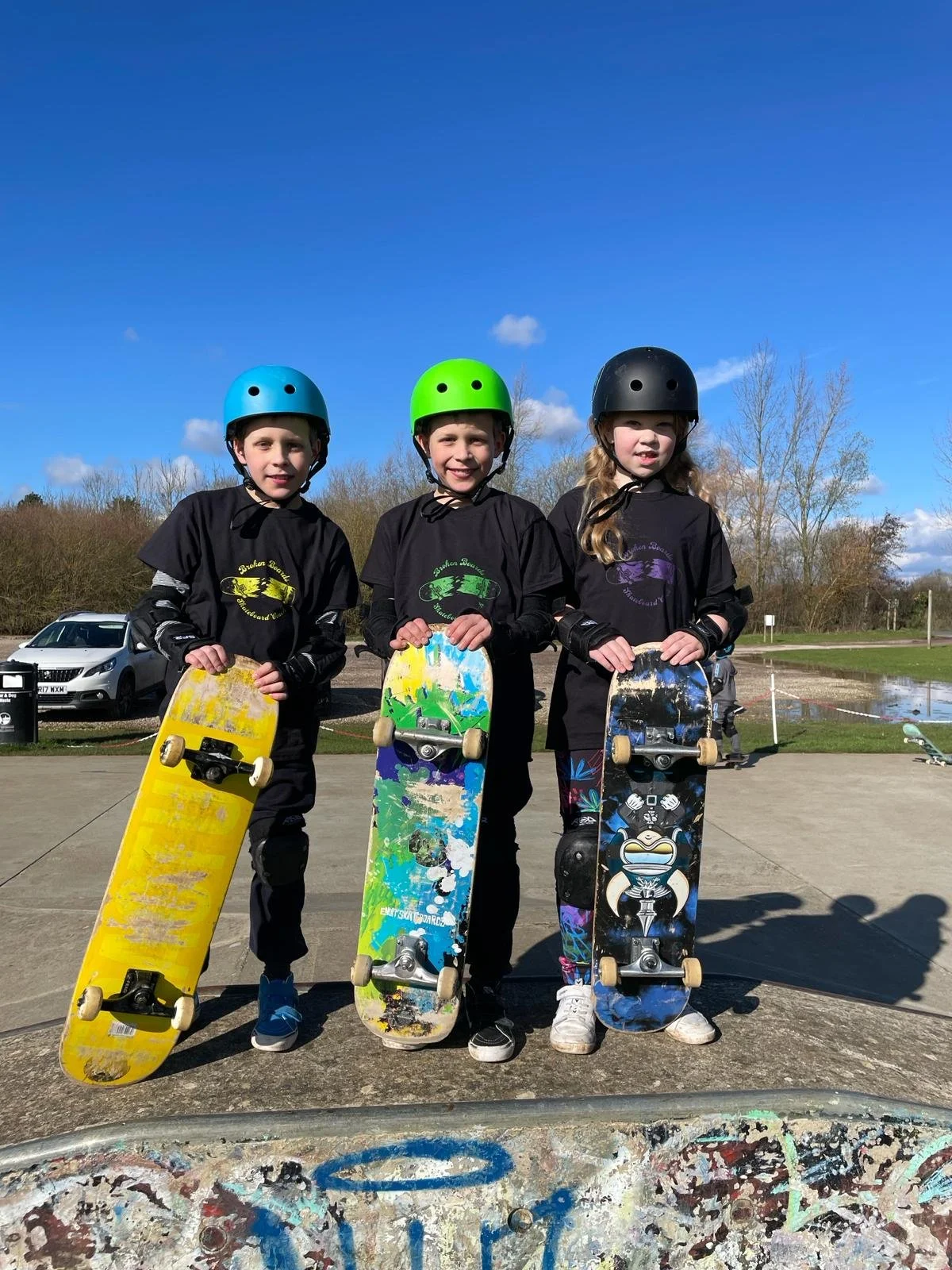 Three young children wearing helmets and protective gear, holding skateboards, standing outdoors on a sunny day.