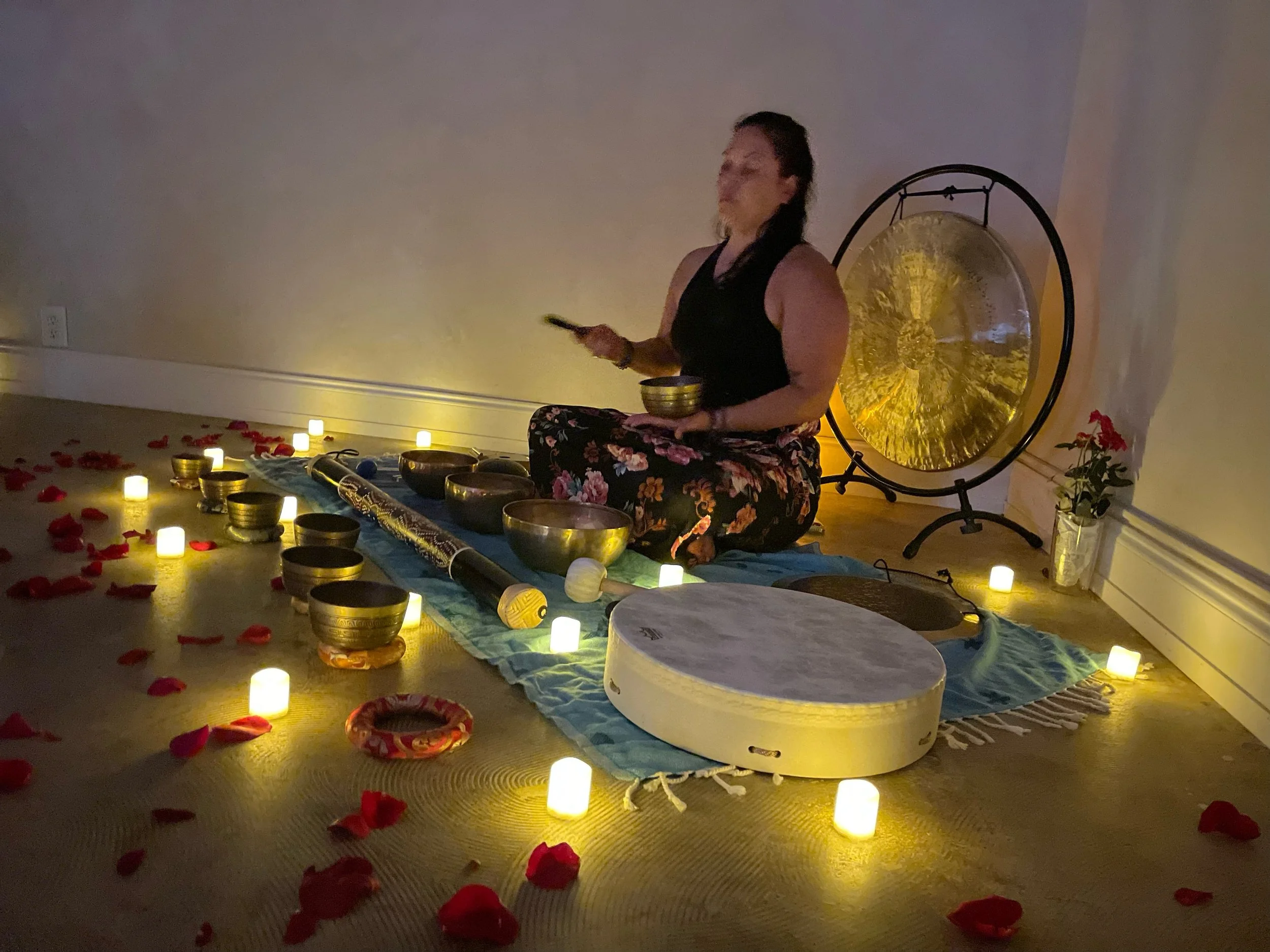 A woman sitting cross-legged with a bowl in hand surrounded by candles and rose petals, playing singing bowls in a dimly lit room with a large gong in the background.