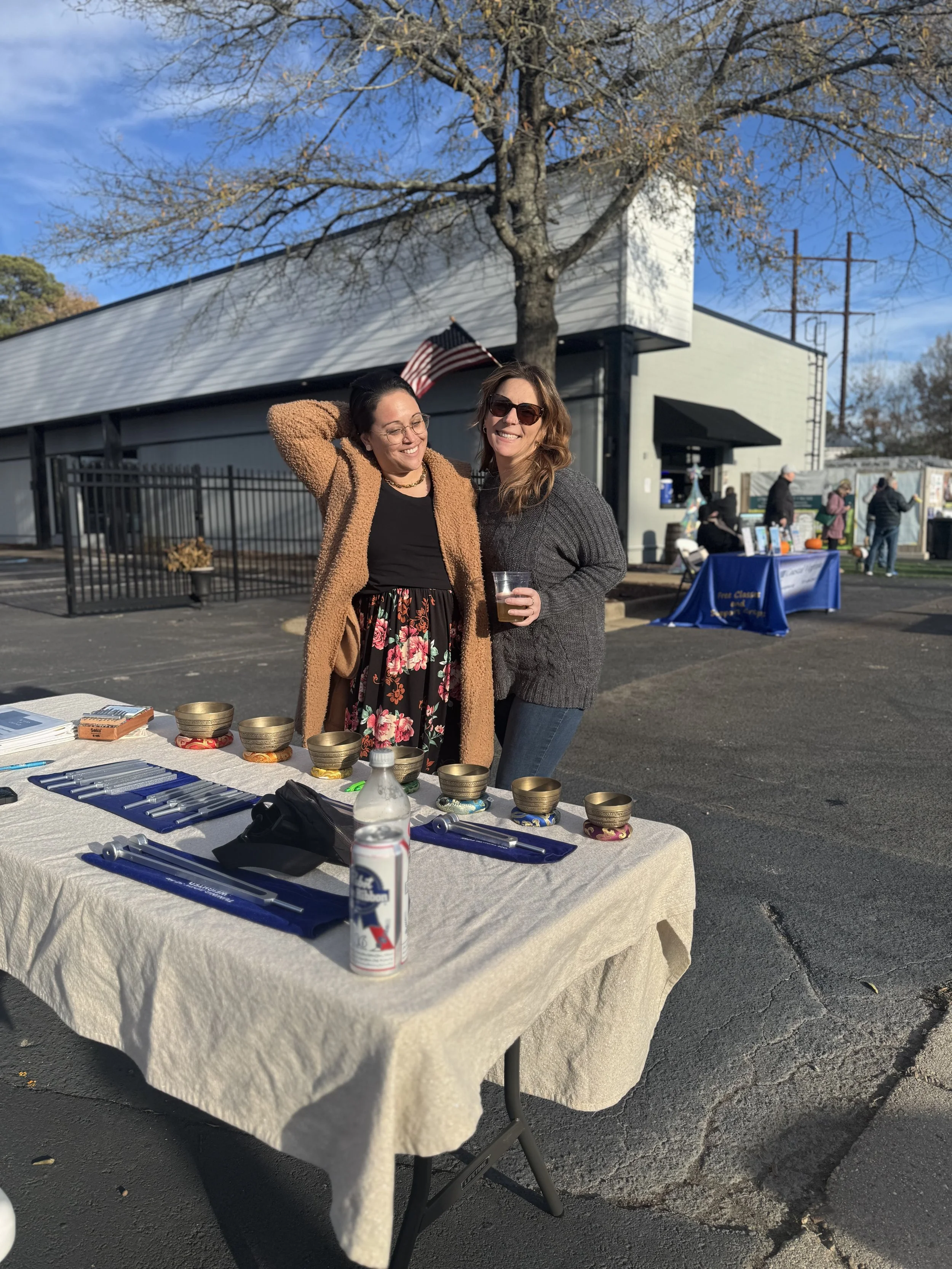 Two women smiling and standing together behind a table displaying singing bowls and writing supplies outdoors. One woman has dark hair, glasses, and is wearing a brown coat, while the other has light brown hair, sunglasses, and a gray sweater. There are trees, a building, and a few people in the background.