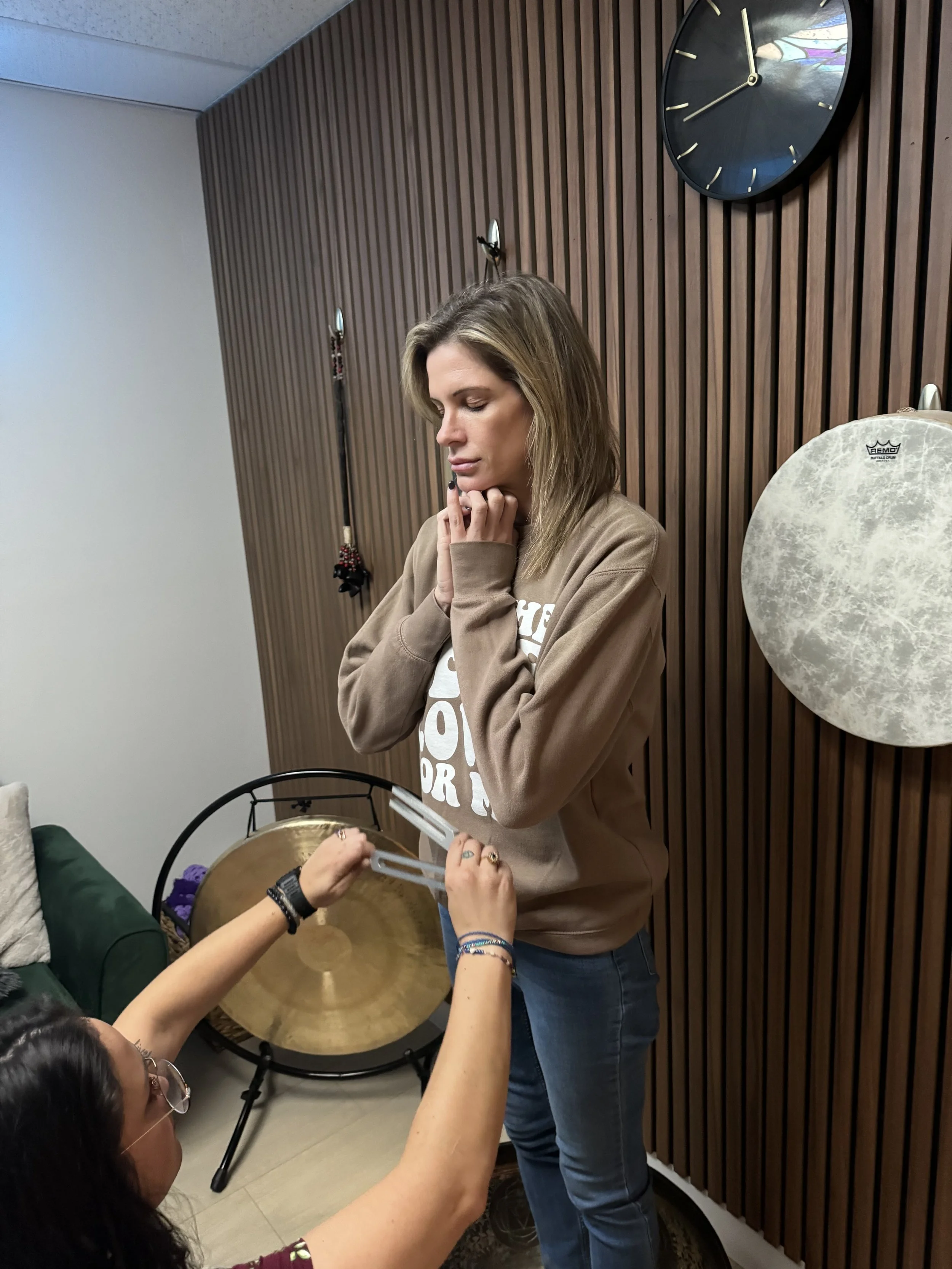 A woman with long blonde hair and wearing a brown hoodie stands in front of a wood-paneled wall with a large round black clock. Multiple people are taking photos of her, some holding a large brass gong.