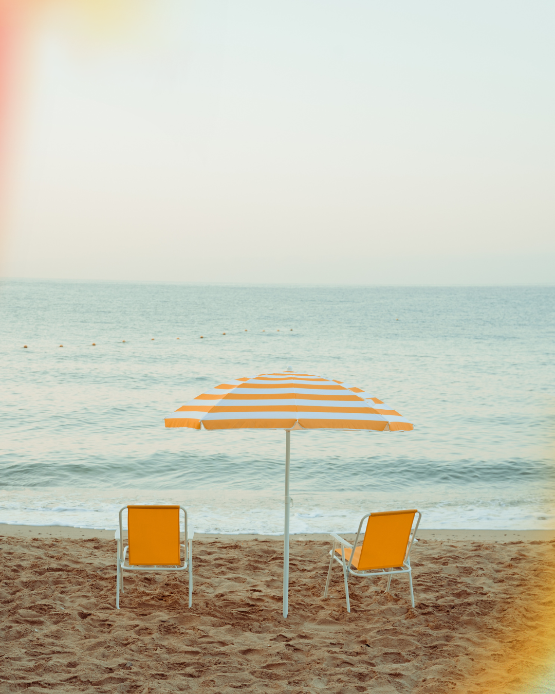 Empty sandy beach with two orange beach chairs and an orange and white striped umbrella facing the ocean, with calm water and a clear sky.
