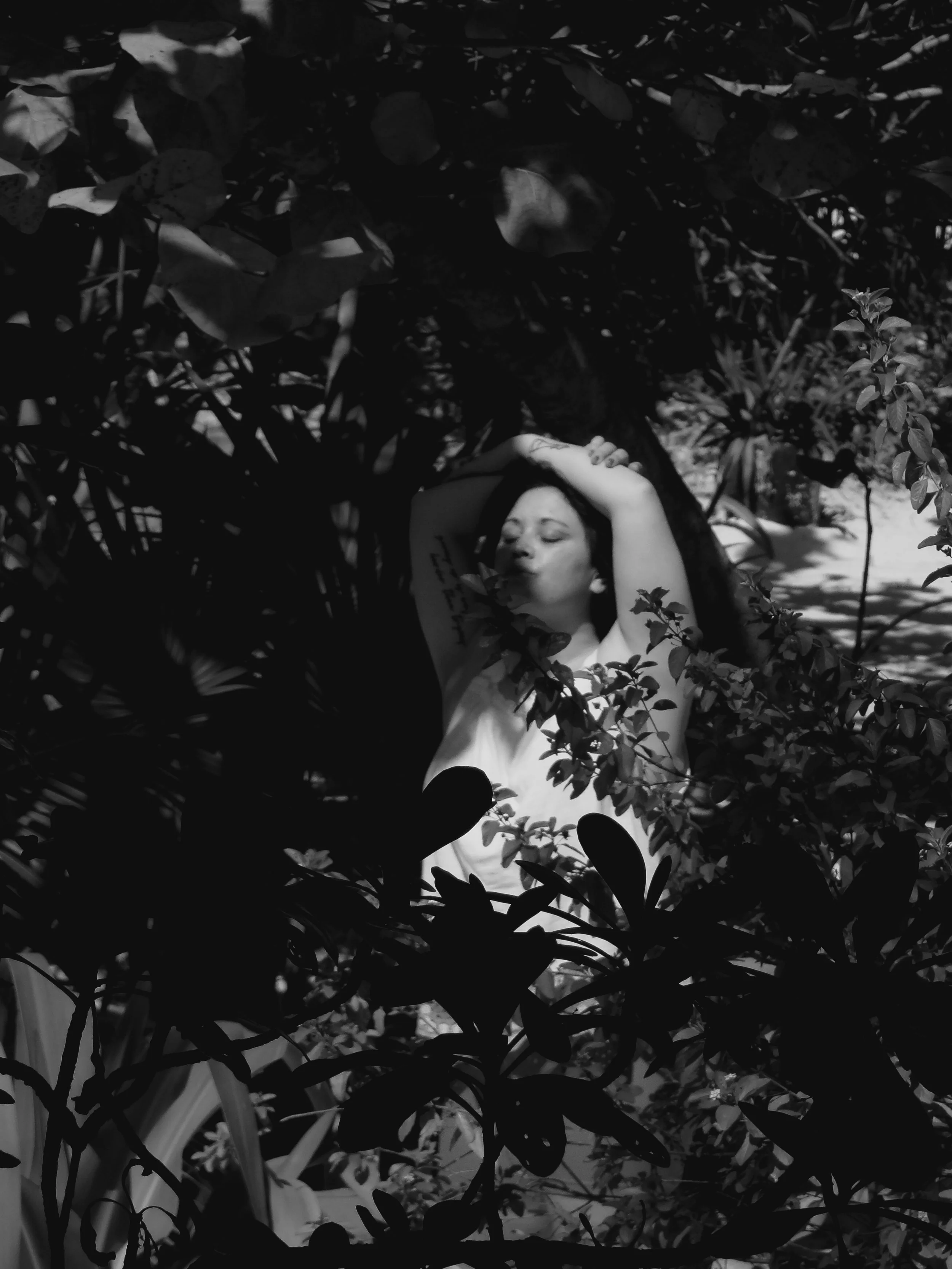 Black and white photo of a woman lying among dense foliage, with her eyes closed and one arm resting on her forehead.