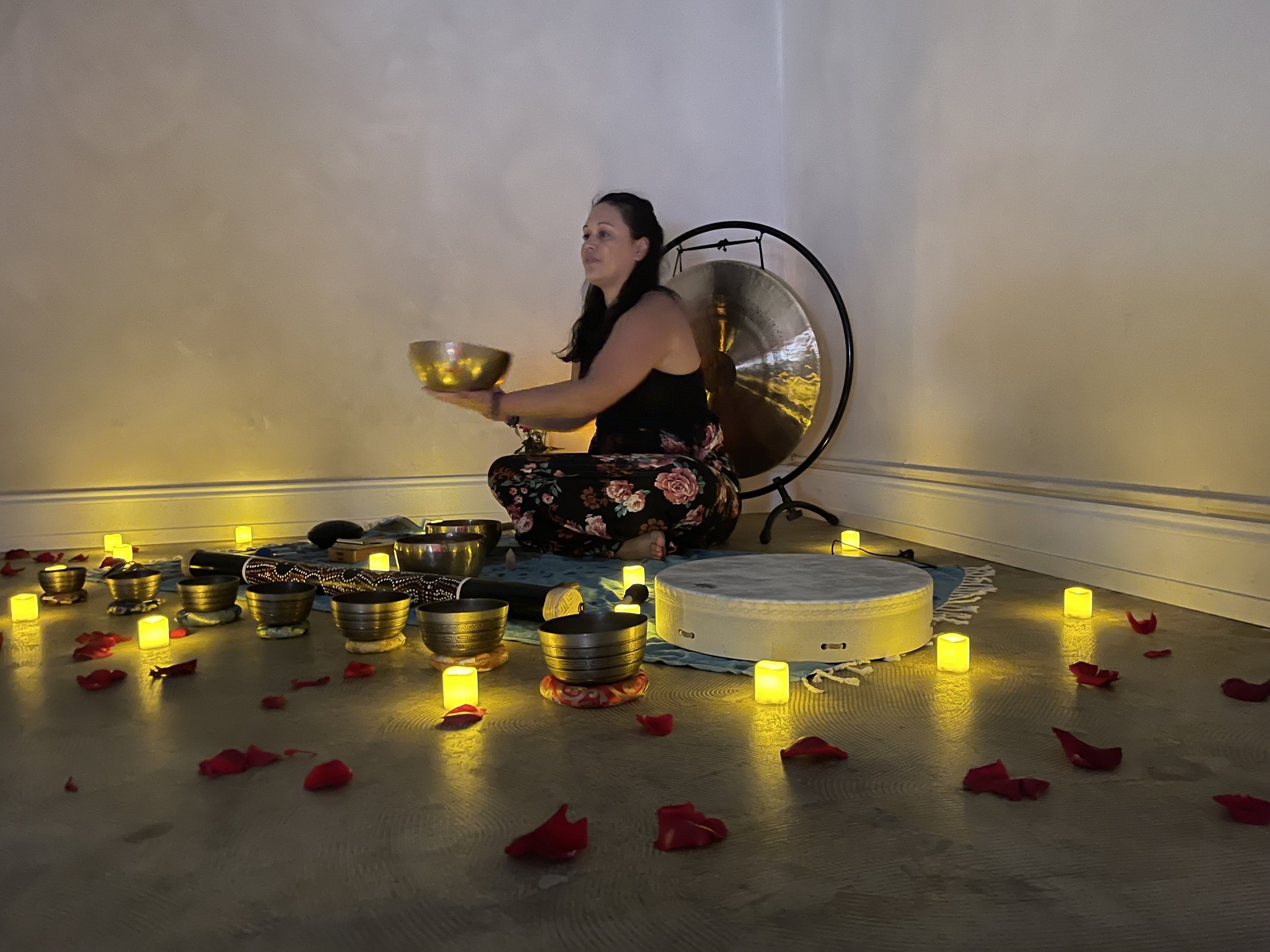 A woman sitting cross-legged on a mat surrounded by lit yellow candles and rose petals, holding a singing bowl, with a large gong behind her, in a calm and serene setting.