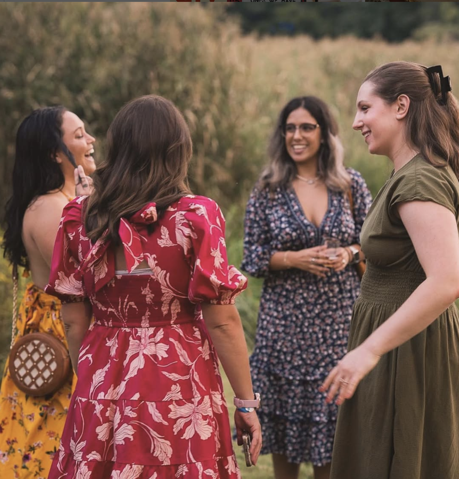 Four women standing outdoors in a field, smiling and engaging in conversation during daytime, with greenery and trees in the background.