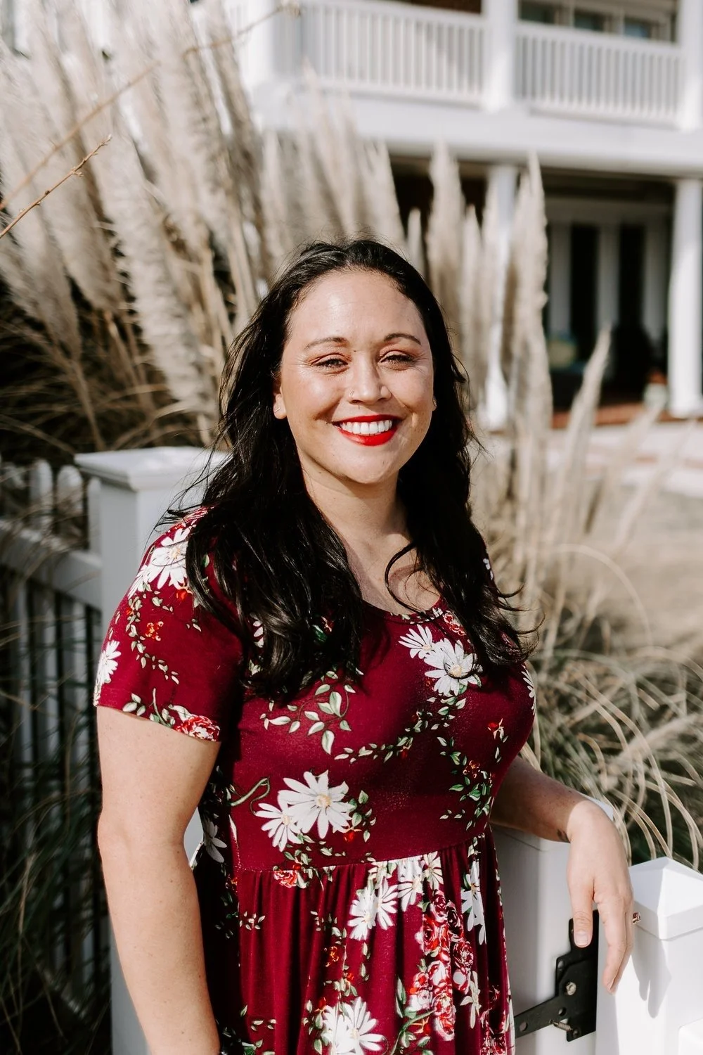 A woman with long dark hair and red lipstick smiling outdoors, wearing a red floral dress, with tall ornamental grass and a building with balconies in the background.