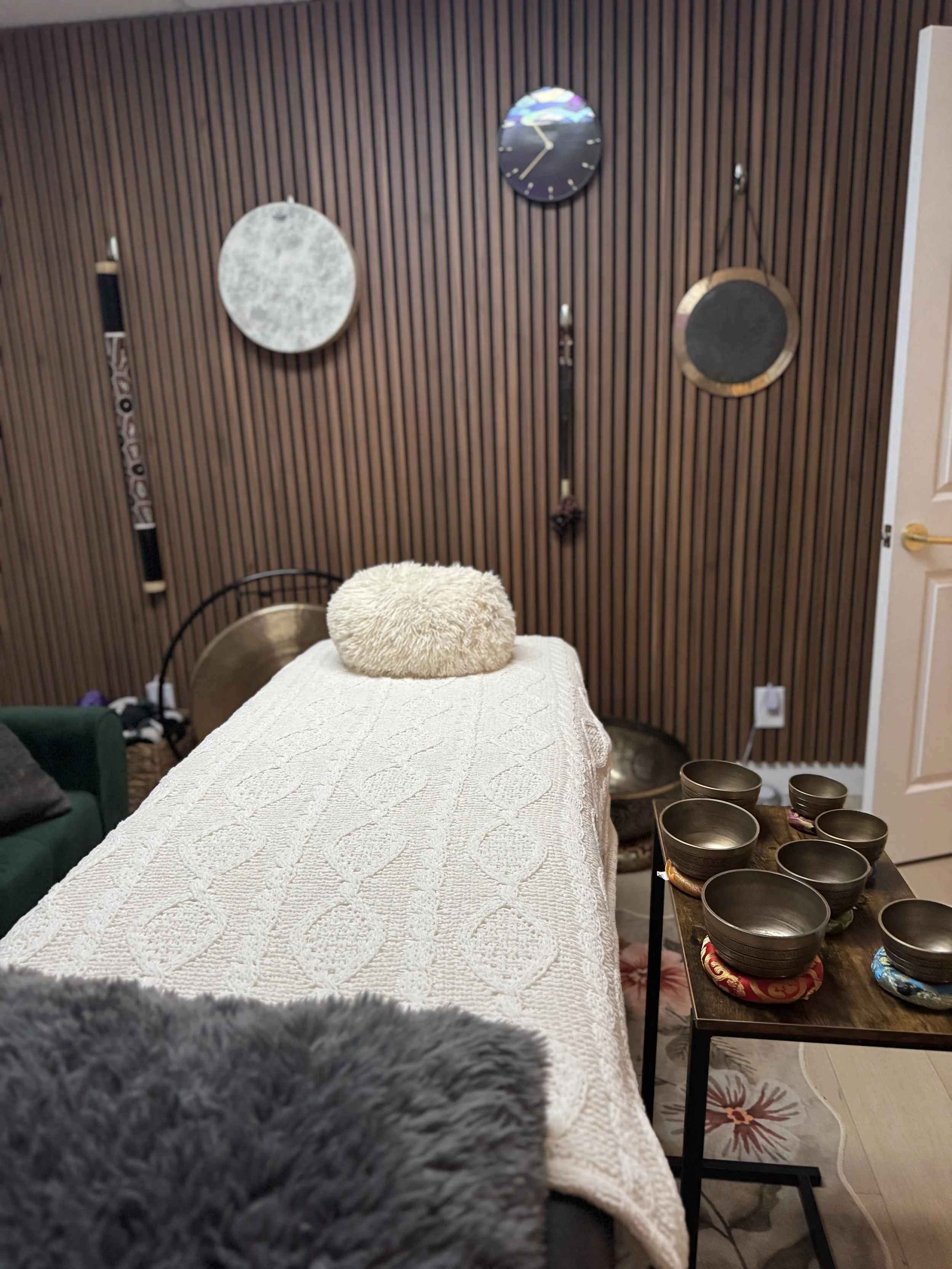 A therapy or massage room with a massage table covered in a white textured blanket, a fluffy round pillow, and a dark wooden side table with black singing bowls. The background has a wooden slat wall with three clocks and a gong.