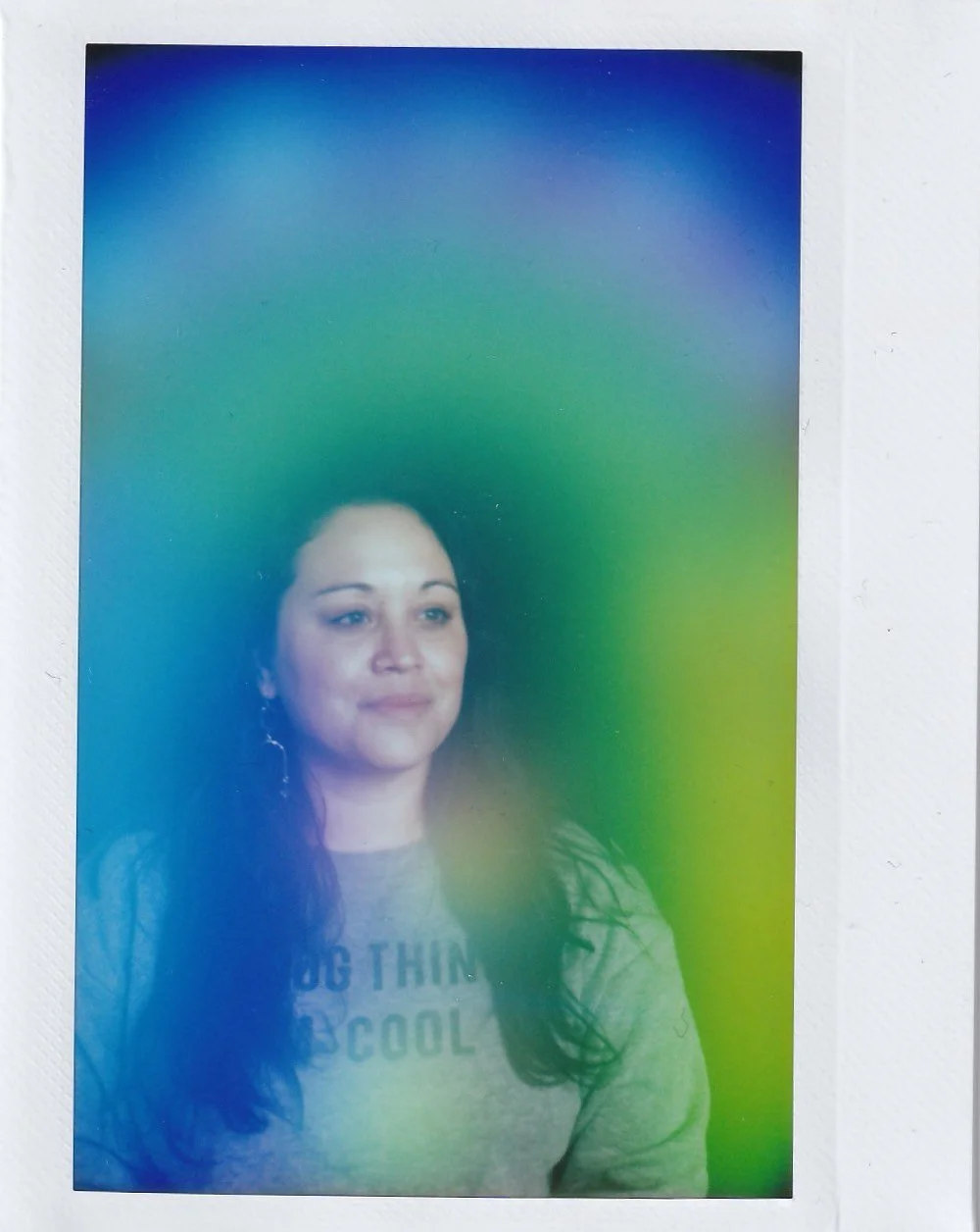 A woman with long dark hair, wearing a grey shirt that says 'DO THINK COOL', stands against a blurred green and blue background with rainbow-like colors.