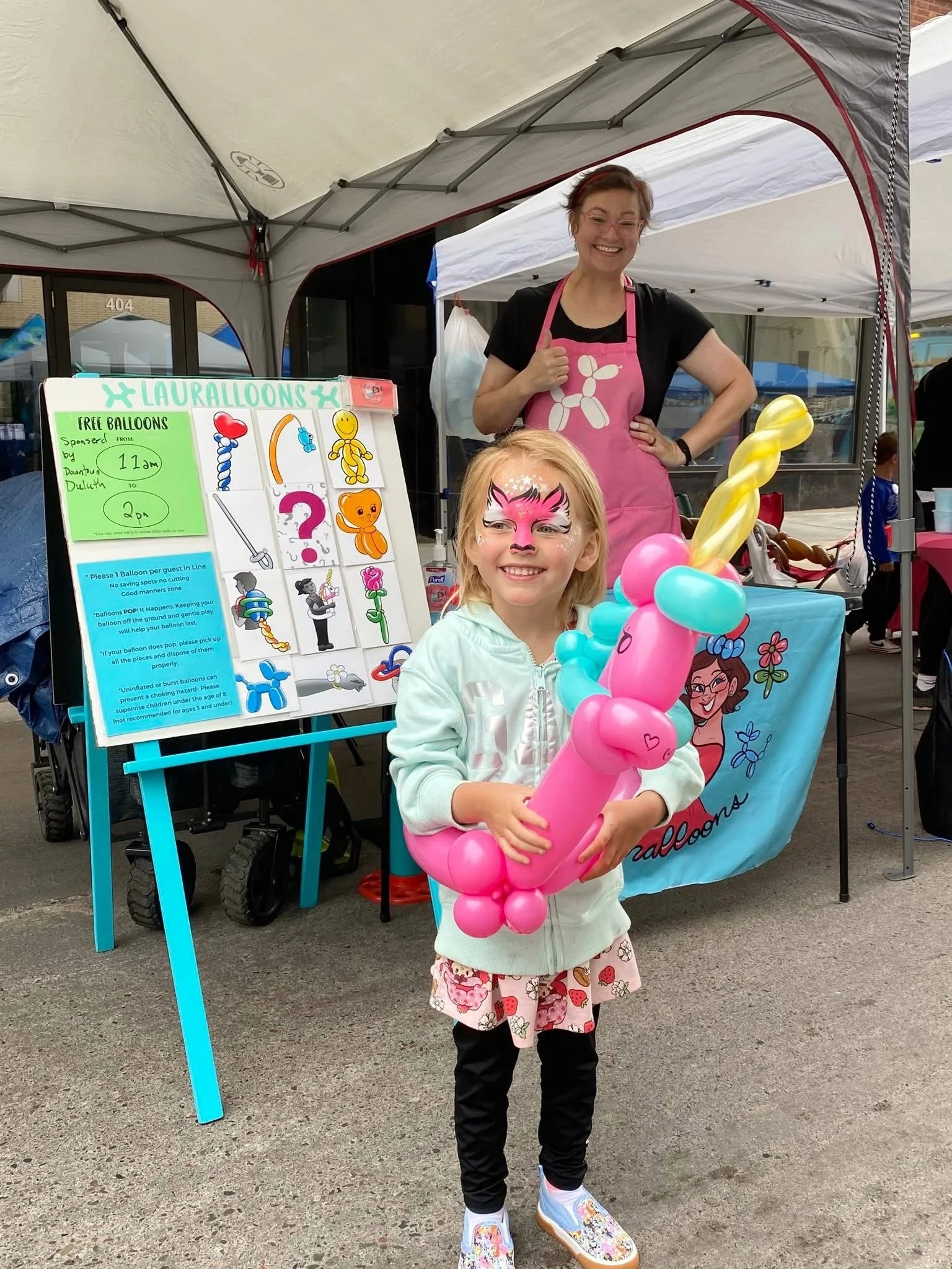 Balloon Artist Laural Schultze, in the background with her twisting set up. Little girl in foreground with facepainting and wearing a balloon unicorn