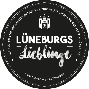 Circular logo with a black background, white text, and a small illustration of a tower at the top. The text reads: 'Mit besten Empfehlungen: Entdecke deine neuen Lieblinge der Region Lüneburg. Lüneburgs Lieblinge. www.lueneburgs-lieblinge.de'.