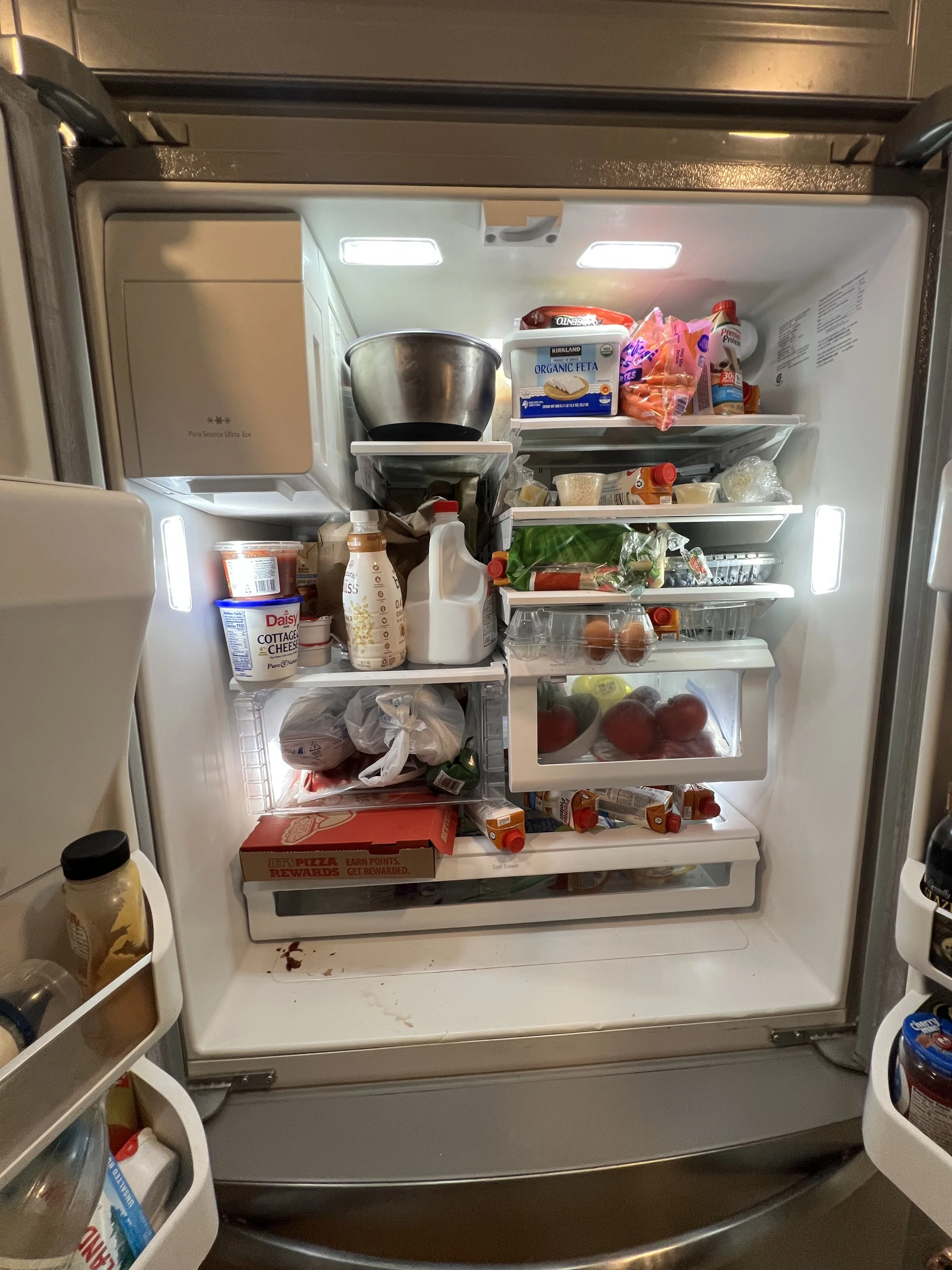 Open refrigerator with various food items including dairy, produce, condiments, and leftovers, with a pizza box at the bottom.
