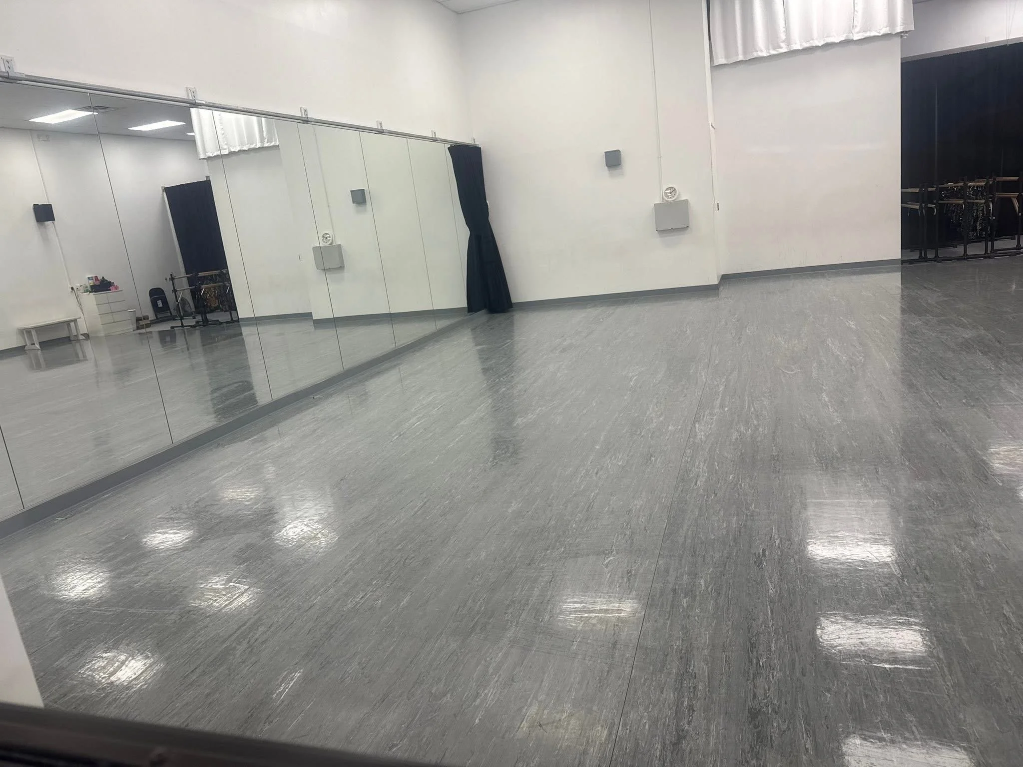 Empty dance studio with light-colored tiled floor, large mirror wall, and white walls. There are black curtains on the side with some equipment and furniture visible in the background.