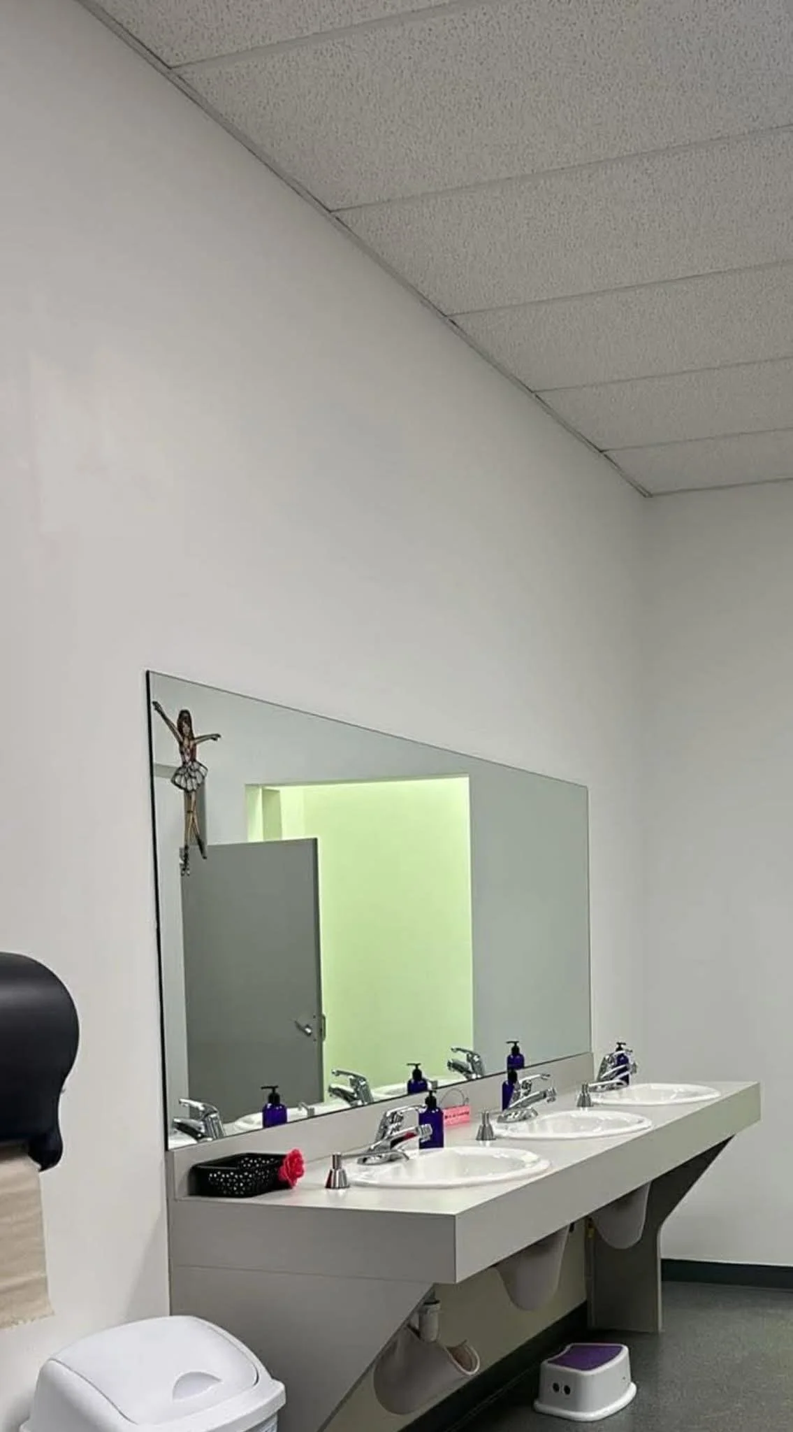 A public restroom with a large mirror, three sinks with soap dispensers, and a small stool underneath. There is a towel dispenser on the left, a trash can beneath the towel dispenser, and a wall decoration of a ballerina hanging on the mirror.