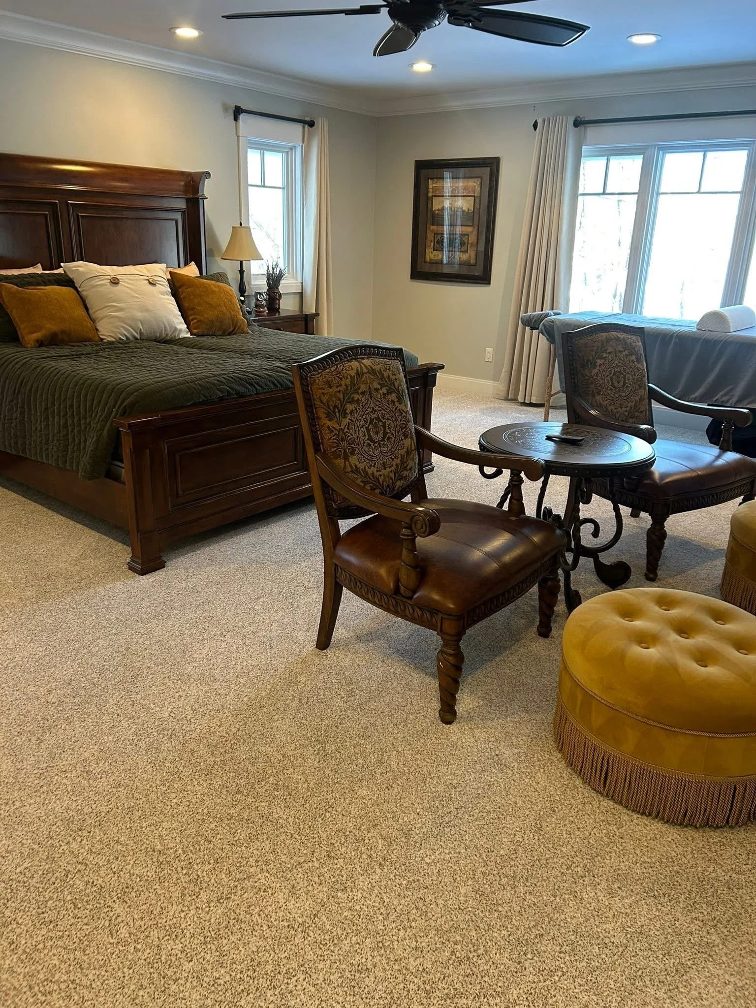 A bedroom with a large wooden bed, two windows with white curtains, a dark wooden nightstand, a lamp, framed artwork on the wall, and a sitting area with a decorative round yellow ottoman, two leather chairs, and a small table.