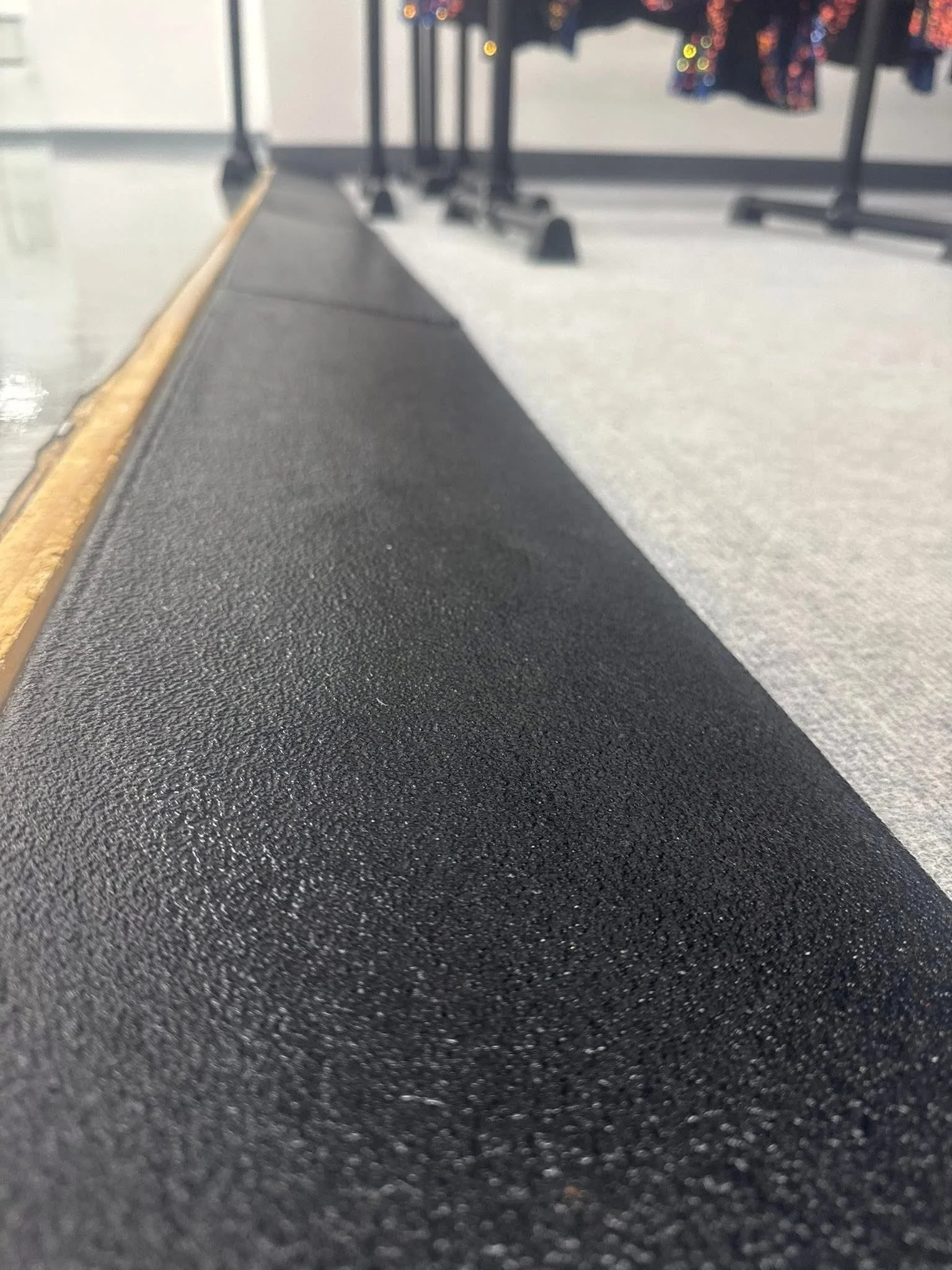 Close-up of a textured black rubber or plastic surface, possibly a sidewalk or mat, with a yellow line on the edge. In the background, there are blurred tables or racks with hanging colorful clothing.