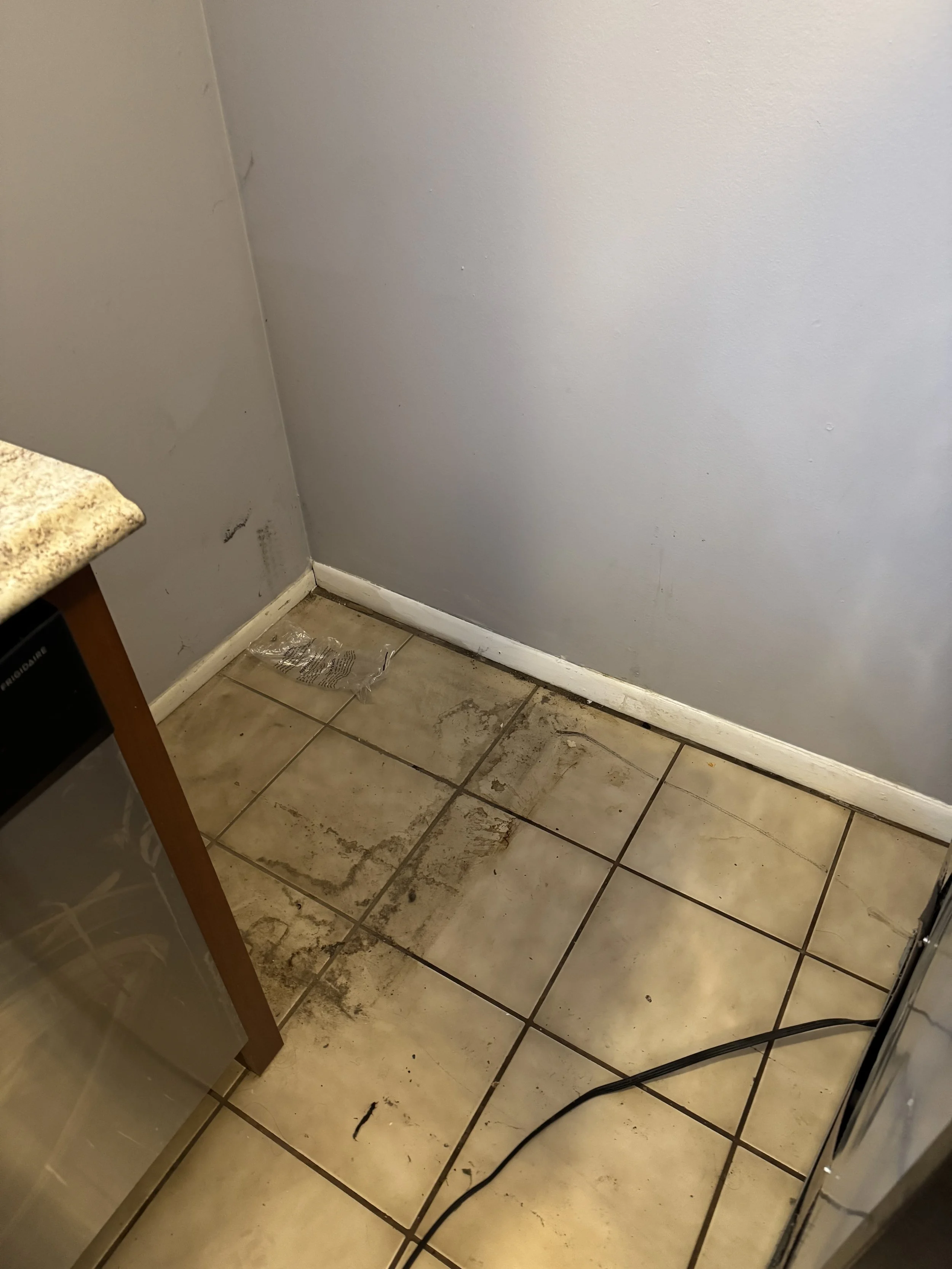 Dirty kitchen corner with stained floor tiles, dirty baseboard, and a cable on the floor.