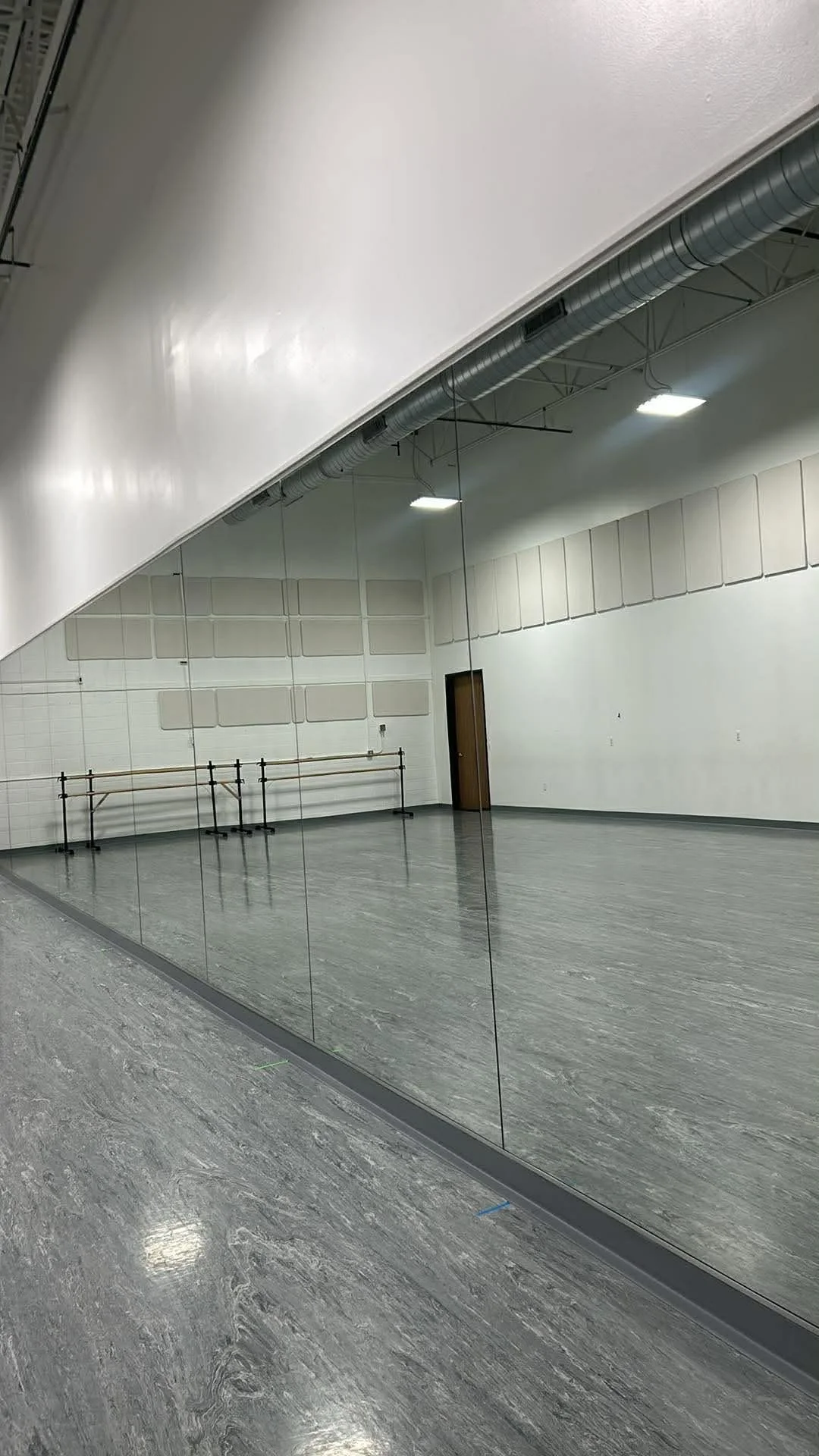 An empty dance or exercise studio with a large mirror on the wall, ballet barres, and grey flooring.