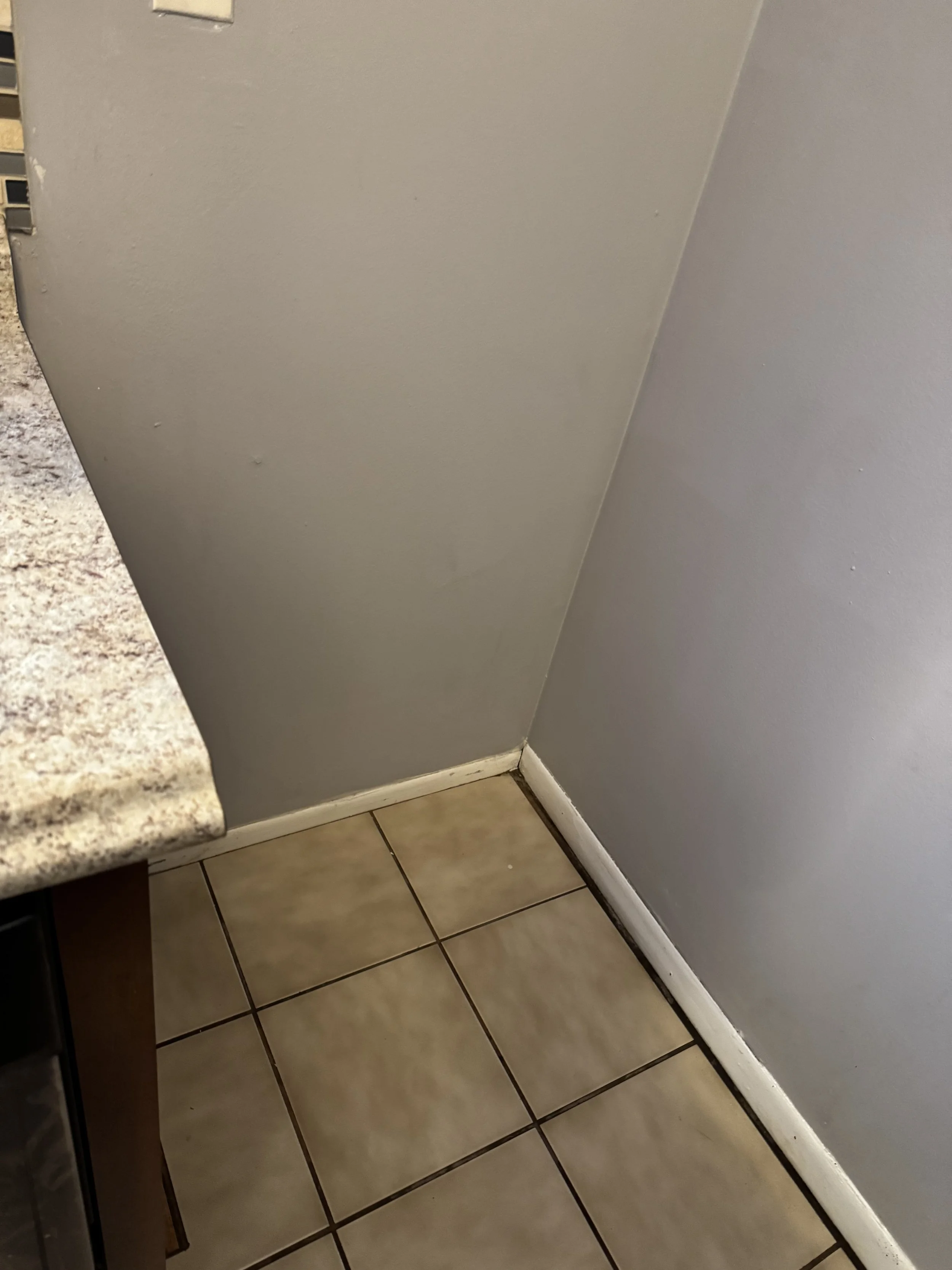 Corner of a room showing a white baseboard, gray painted walls, and beige tiled flooring.