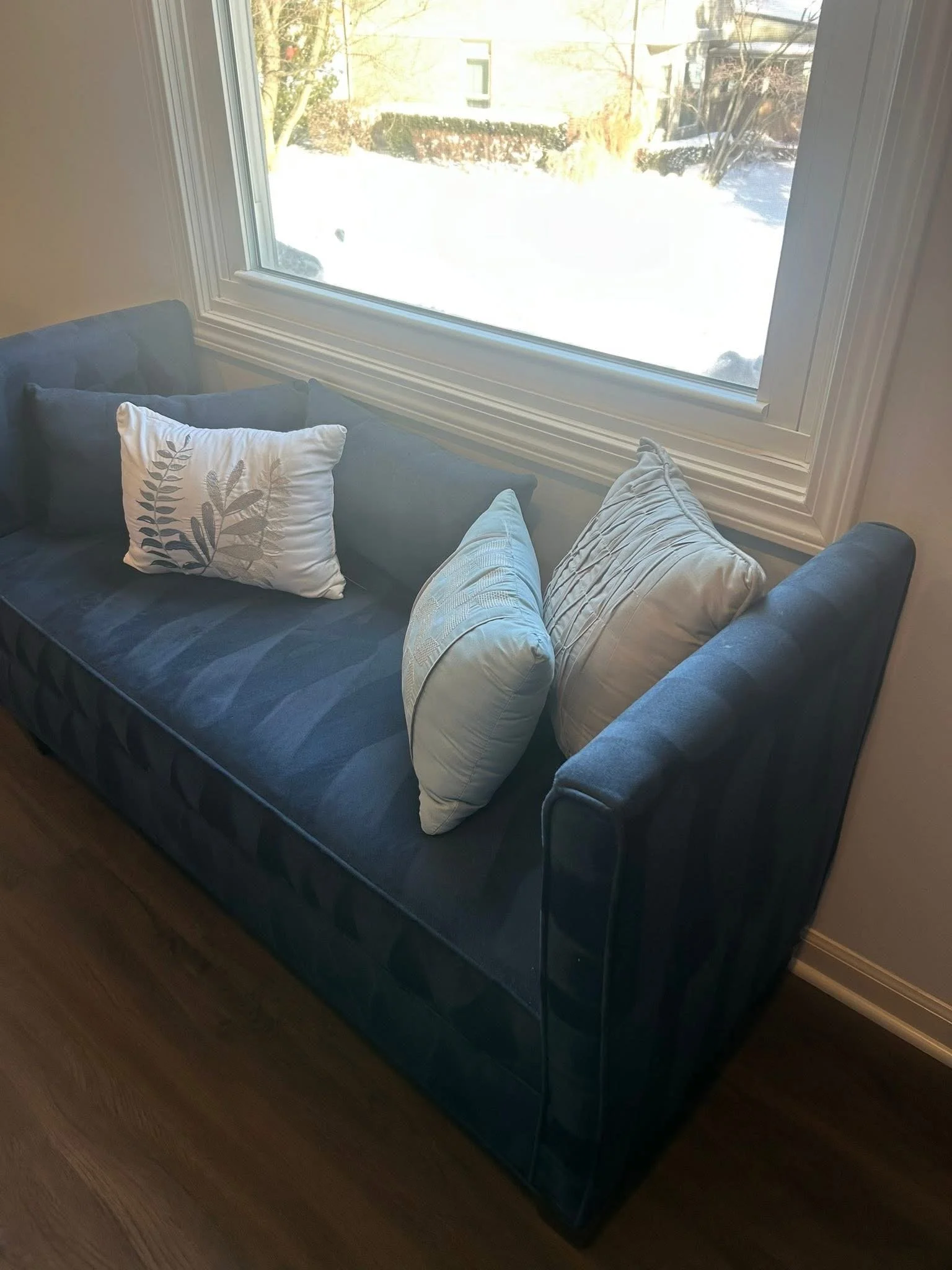 Blue sofa with three decorative pillows next to a large window showing a snowy outdoor scene.