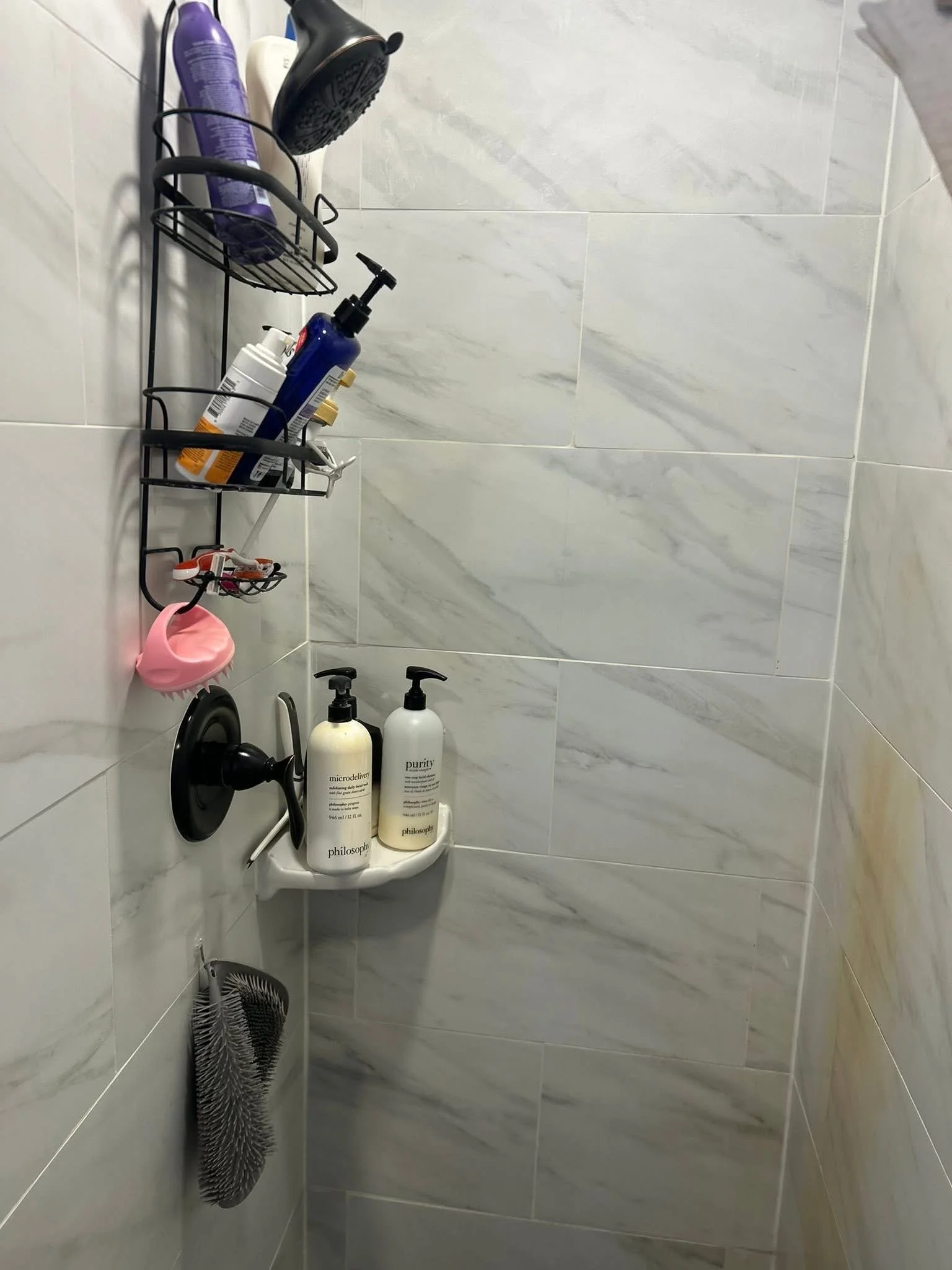 A tiled shower corner with a black wall-mounted showerhead, wire shelf holding bottles of shampoo, conditioner, and body wash, a pink shower scrub, a black hairbrush, and two bottles of Philosophy brand lotion on a small white shelf.
