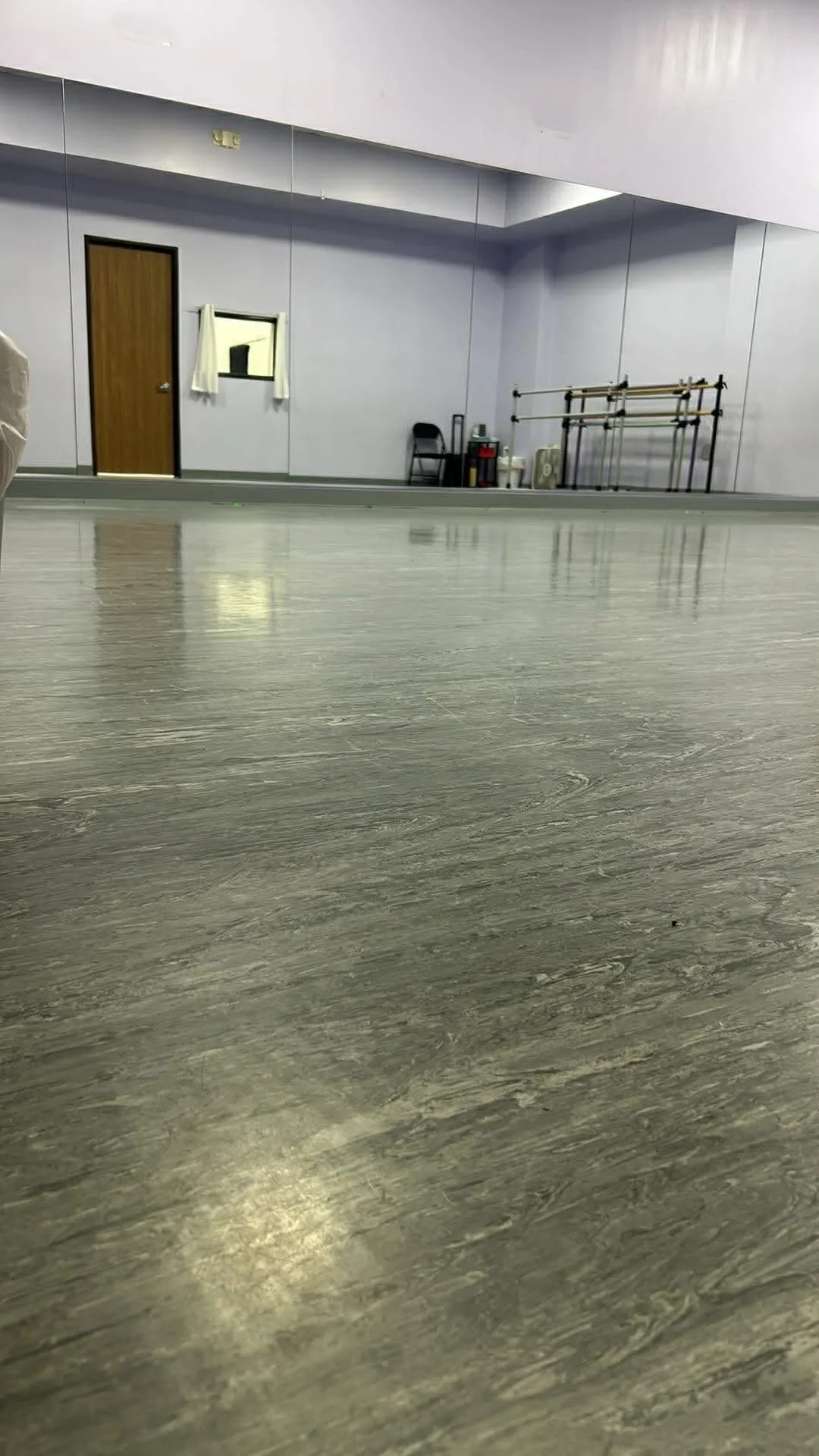An empty dance studio with a wood-finished floor, gray walls, a door, a small window with curtains, a chair, and exercise equipment in the background.
