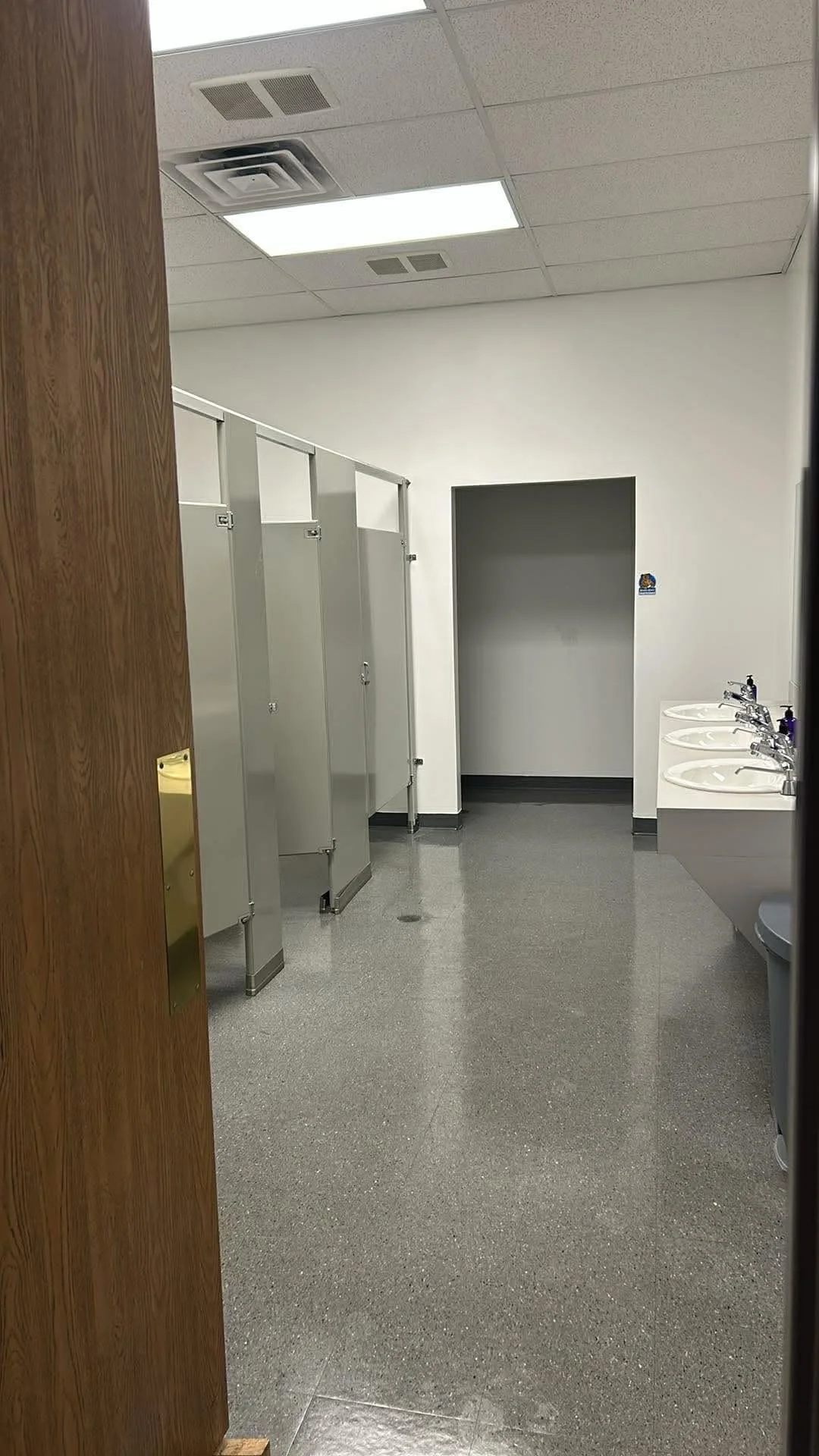 A public restroom with three gray stall doors, a doorway leading to an empty room, and a row of three white sinks with silver faucets and purple soap dispensers. The floor is gray, and the ceiling has white tiles with fluorescent lighting.