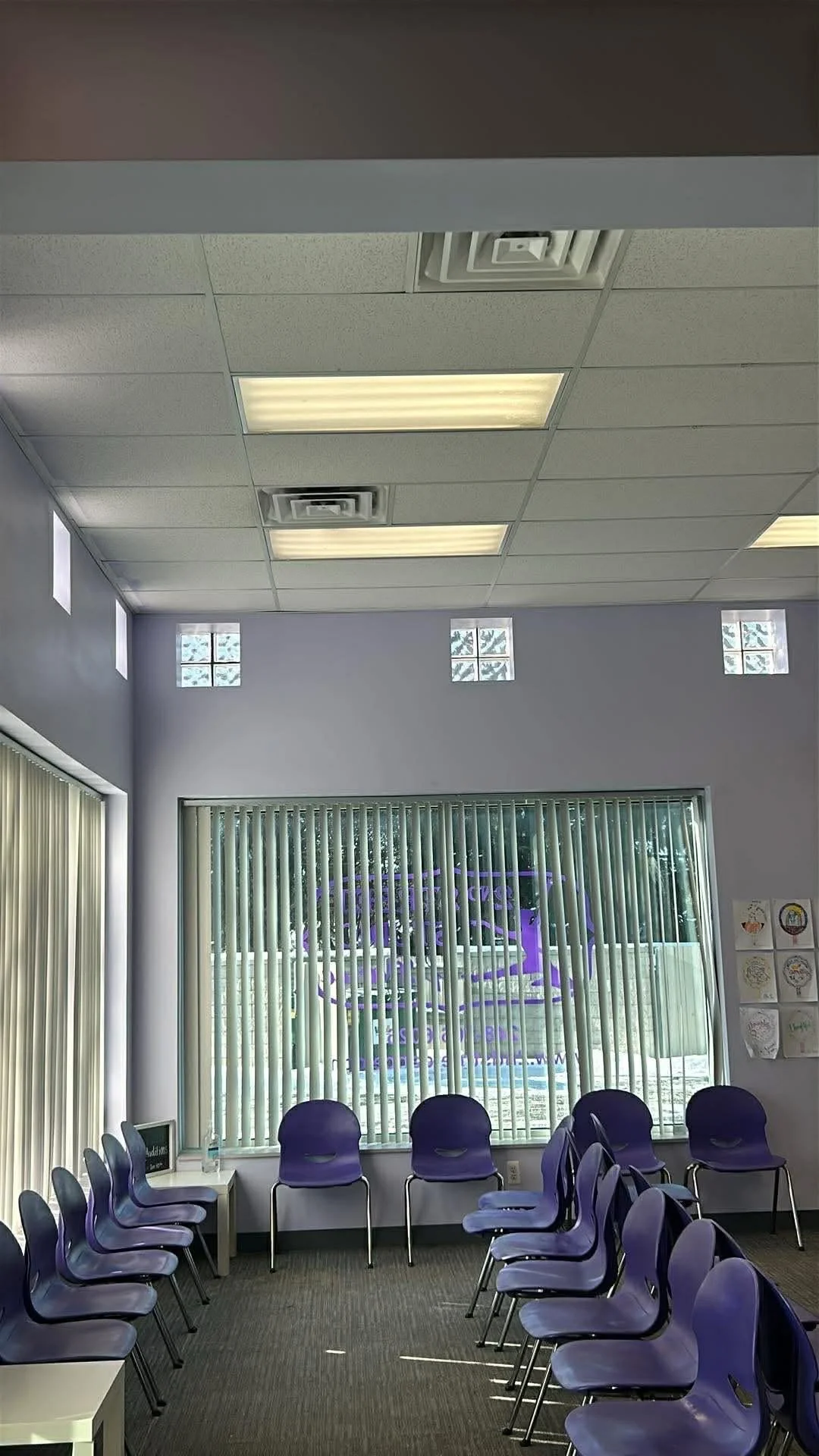 A waiting room with purple chairs arranged along the walls and in the center. Large window with vertical blinds, some small window openings at the top of the walls, and ceiling with ceiling tiles and fluorescent lights.