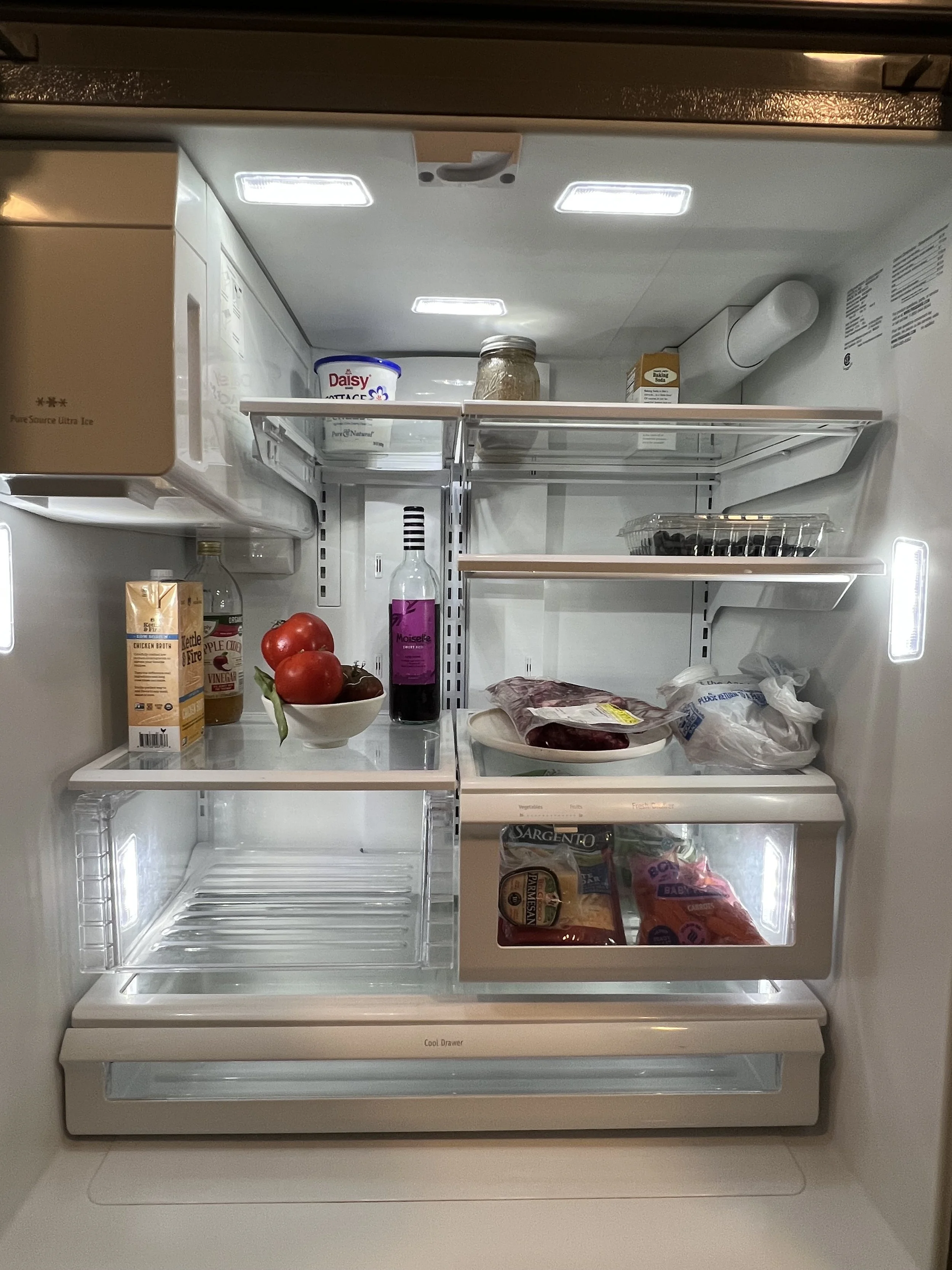 Open refrigerator door showing various food items including tomatoes, a bottle of wine, packaged meat, frozen vegetables, and condiments.