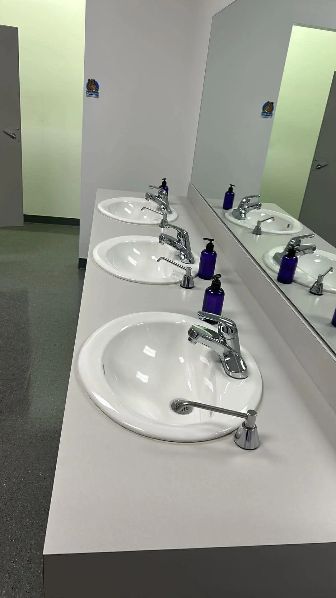 Public restroom with three white sinks, each with a silver faucet, purple soap dispensers, and a large mirror reflecting the sinks and soap dispensers.