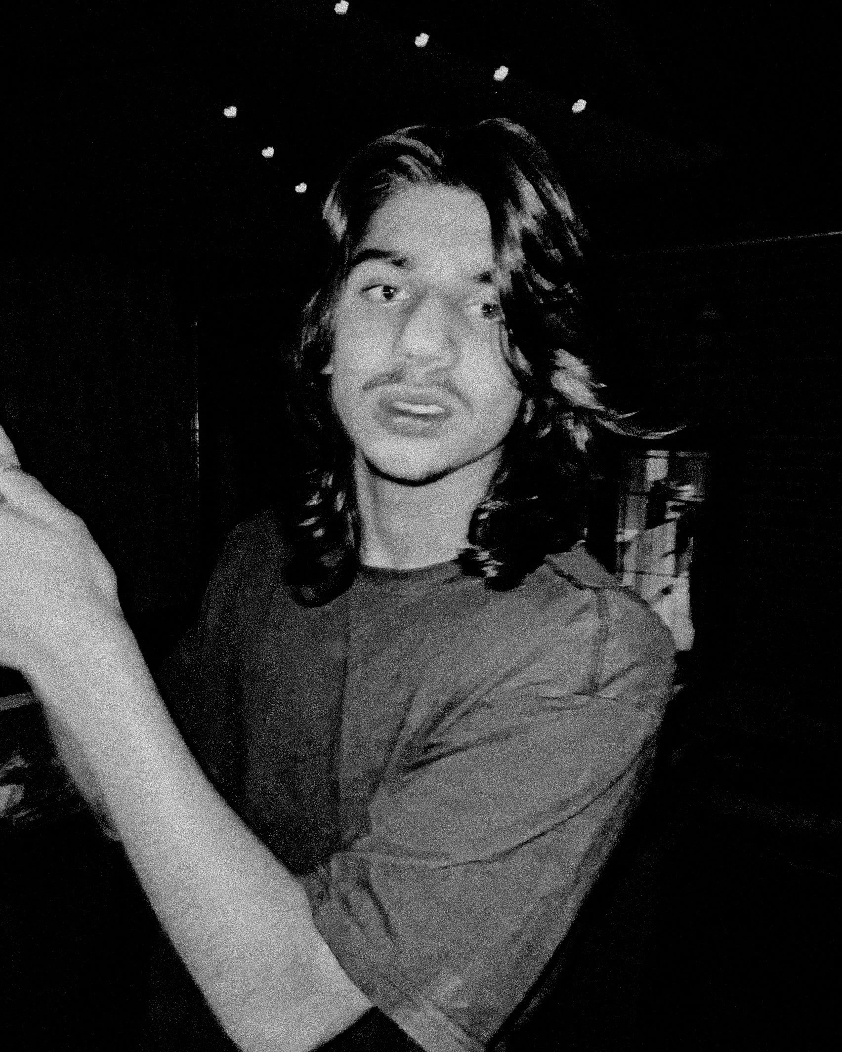 A black-and-white photo of a young man with long wavy hair, smiling slightly, in a dimly lit room.