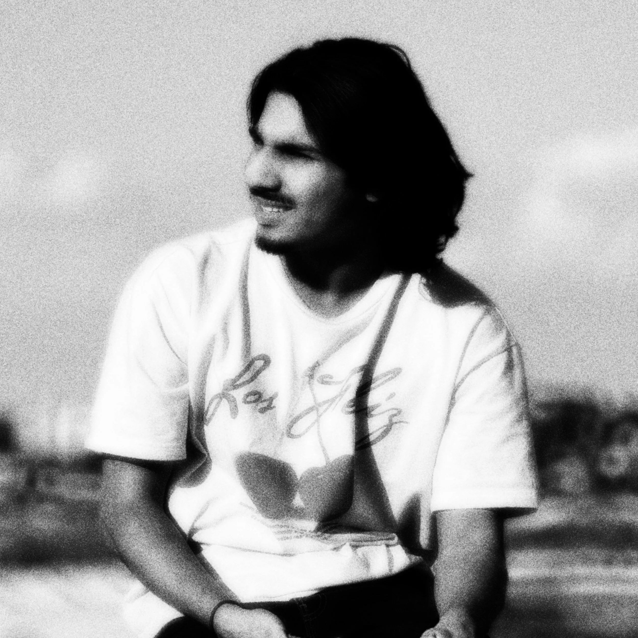 A black-and-white photo of a young man with long wavy hair, smiling slightly, in a dimly lit room.
