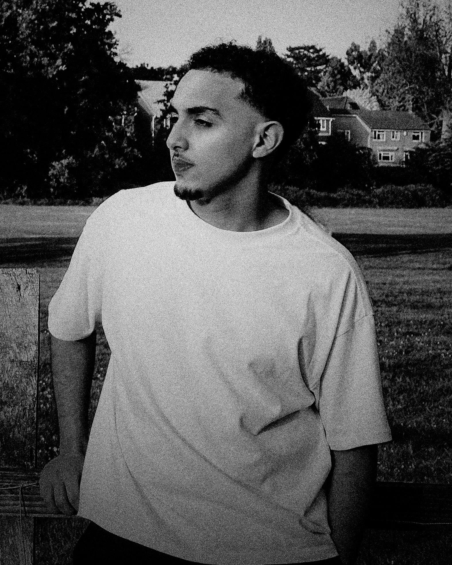 A young man with a short hairstyle, facial hair, and a goatee, standing outdoors by a wooden post. He is wearing a loose white T-shirt, looking to his left, with trees and residential houses in the background.